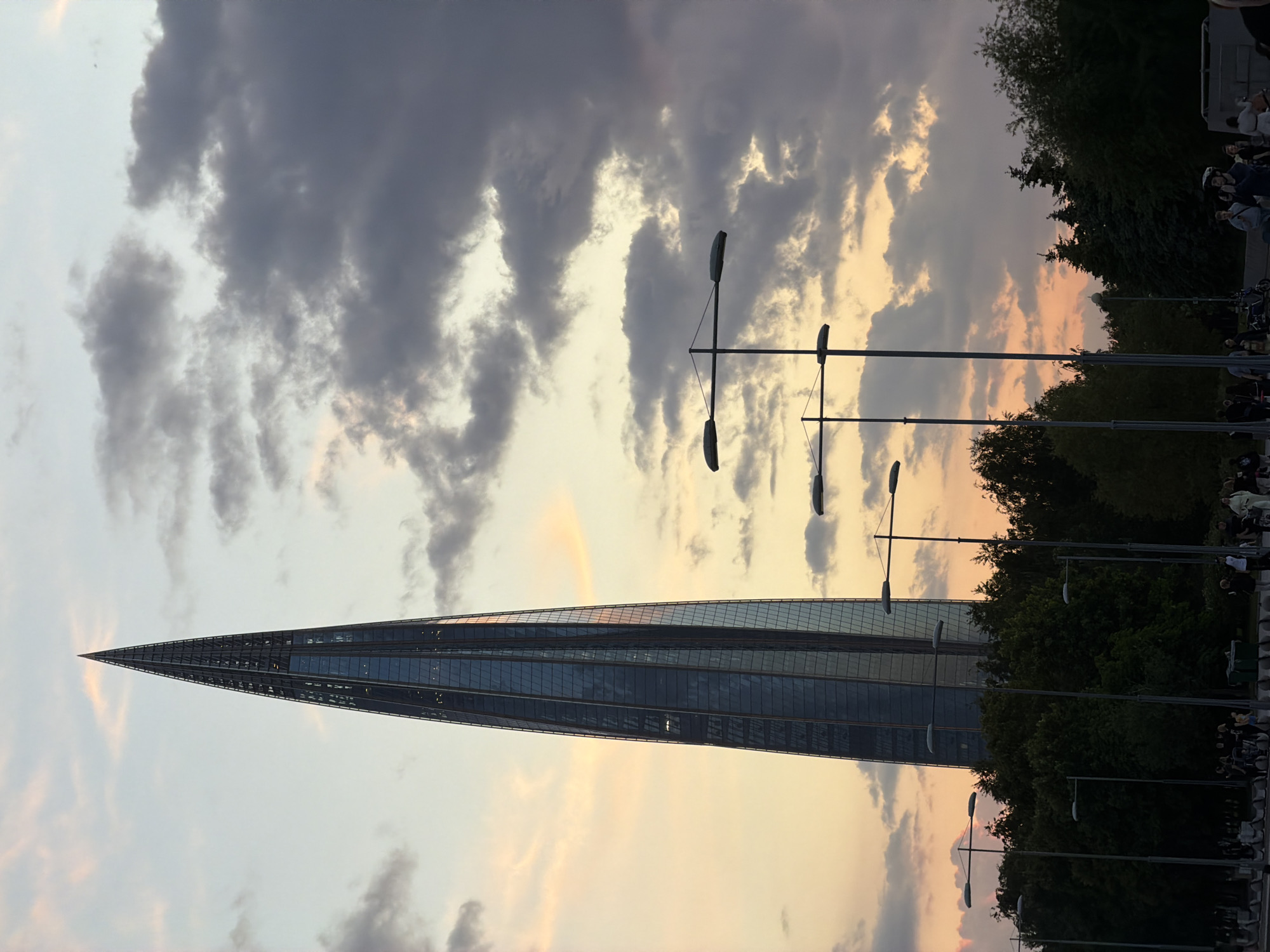 The image displays a prominent, slender, and modern skyscraper, identifiable as the Lakhta Center, rising towards the sky from the lower left portion of the frame in Saint Petersburg, Russia. The building features a distinctive tapering and twisting glass facade, reflecting the surrounding light.
In the foreground and mid-ground, several multi-armed streetlights stand vertically, their dark silhouettes contrasting against the sky. Dark green, dense trees line the right side of the image, obscuring parts of the view. On the far right, behind the trees and streetlights, a group of people is visible, some standing, some seated, suggesting a public outdoor area like a park or promenade. No specific activity or event is clearly discernable among them.
The sky is a notable feature, presenting a dramatic mix of dark grey, voluminous clouds interspersed with lighter, warm hues of yellow, orange, and soft pink. This suggests the time of day is either late afternoon or early evening, likely around sunset or sunrise, creating a captivating backdrop for the skyscraper. The weather appears overcast with significant cloud cover, yet pockets of softer light break through.
No visible text is present in the image. The scene captures a blend of contemporary architecture and natural elements under a dynamic, color-rich sky.