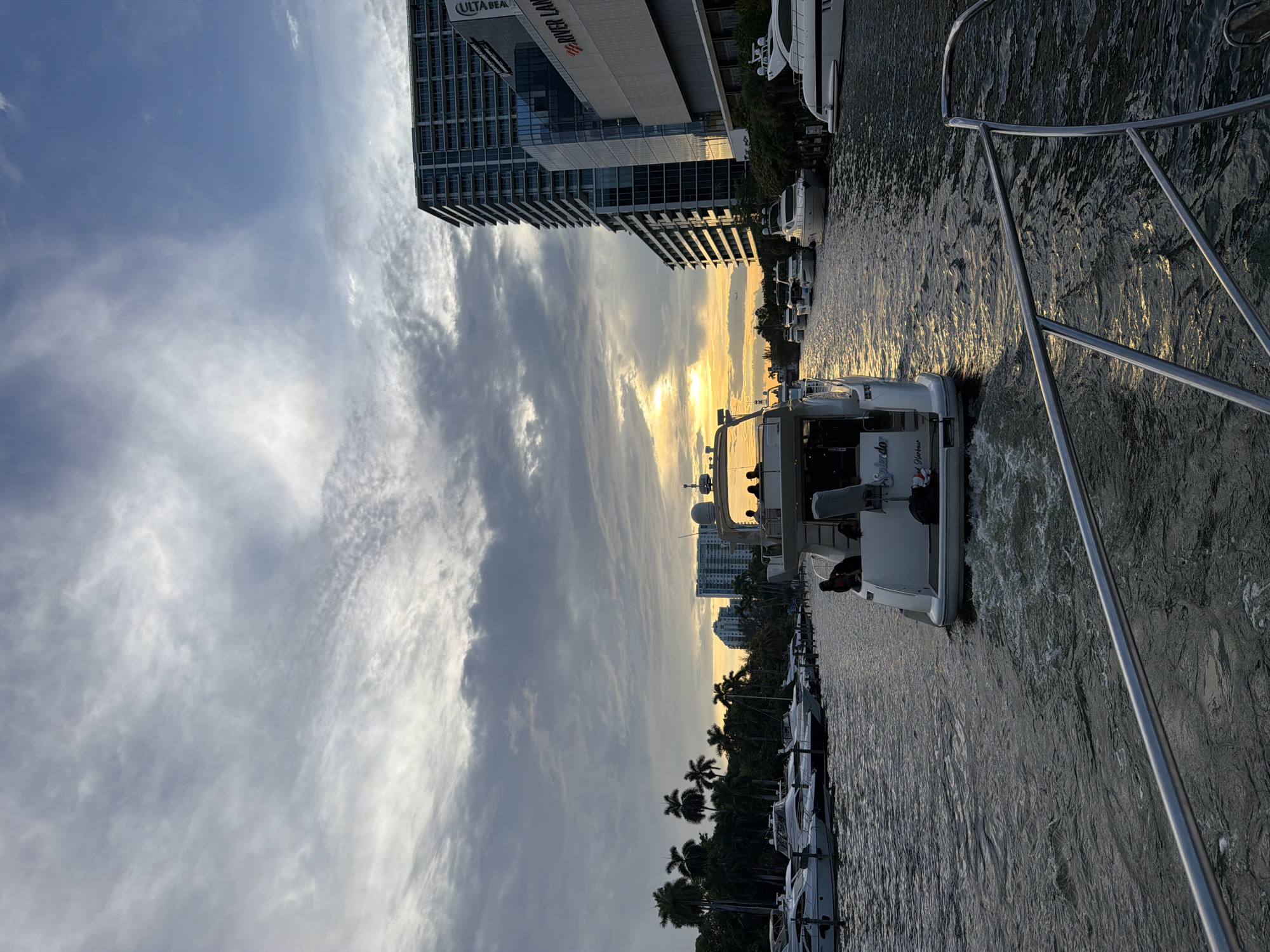 This is a vertical, eye-level shot of a waterway in Miami, Florida, USA, with a fleet of yachts. The sky is cloudy with the sun setting, casting a warm glow on the scene. The water is dark and choppy, reflecting the sky and the surrounding buildings. The boats are lined up along the sides of the waterway, and several people are visible on the decks of the yachts. The buildings in the background are modern and tall, typical of a city skyline. The overall mood is peaceful and serene, with a hint of luxury associated with yachting.