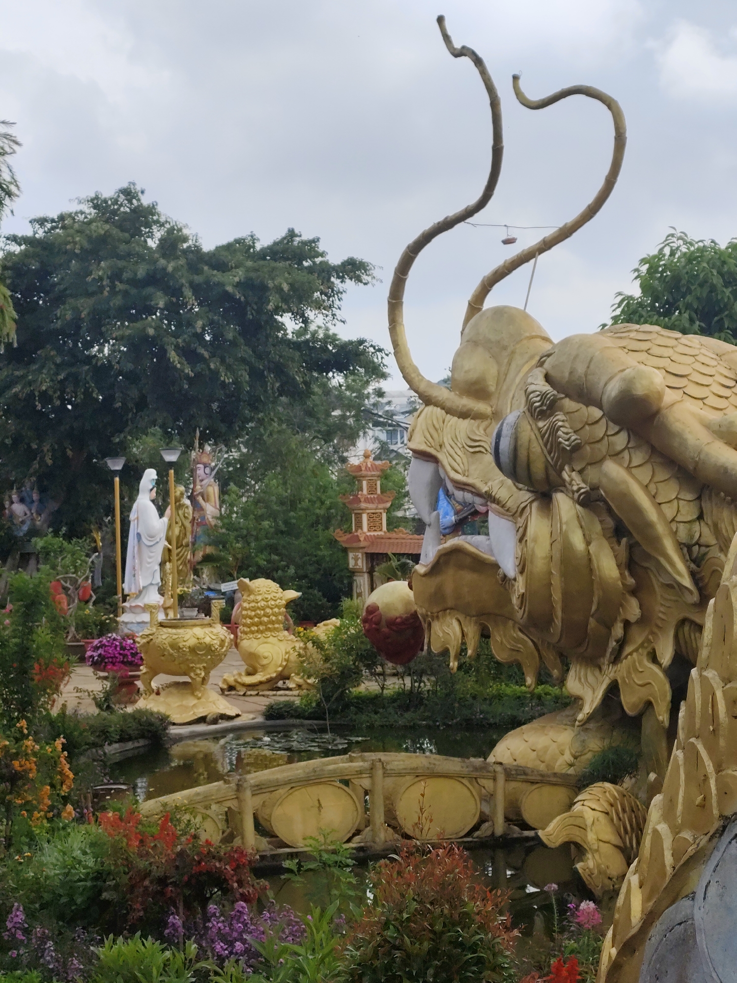 The image captures a scene in the city of Cam Ly - Đà Lạt, Vietnam, featuring a large, golden dragon statue dominating the right side of the frame. The dragon's head, with its prominent horns, scales, and open mouth, is in sharp focus. A bridge crosses a small pond beneath the dragon statue, around which there are colorful flowers and various green plants. In the background, a golden lion statue sits near a white statue of a woman in a flowing robe, with a pagoda-style building visible further back. A large tree with dense foliage occupies the upper-left portion of the image, and the sky is overcast.