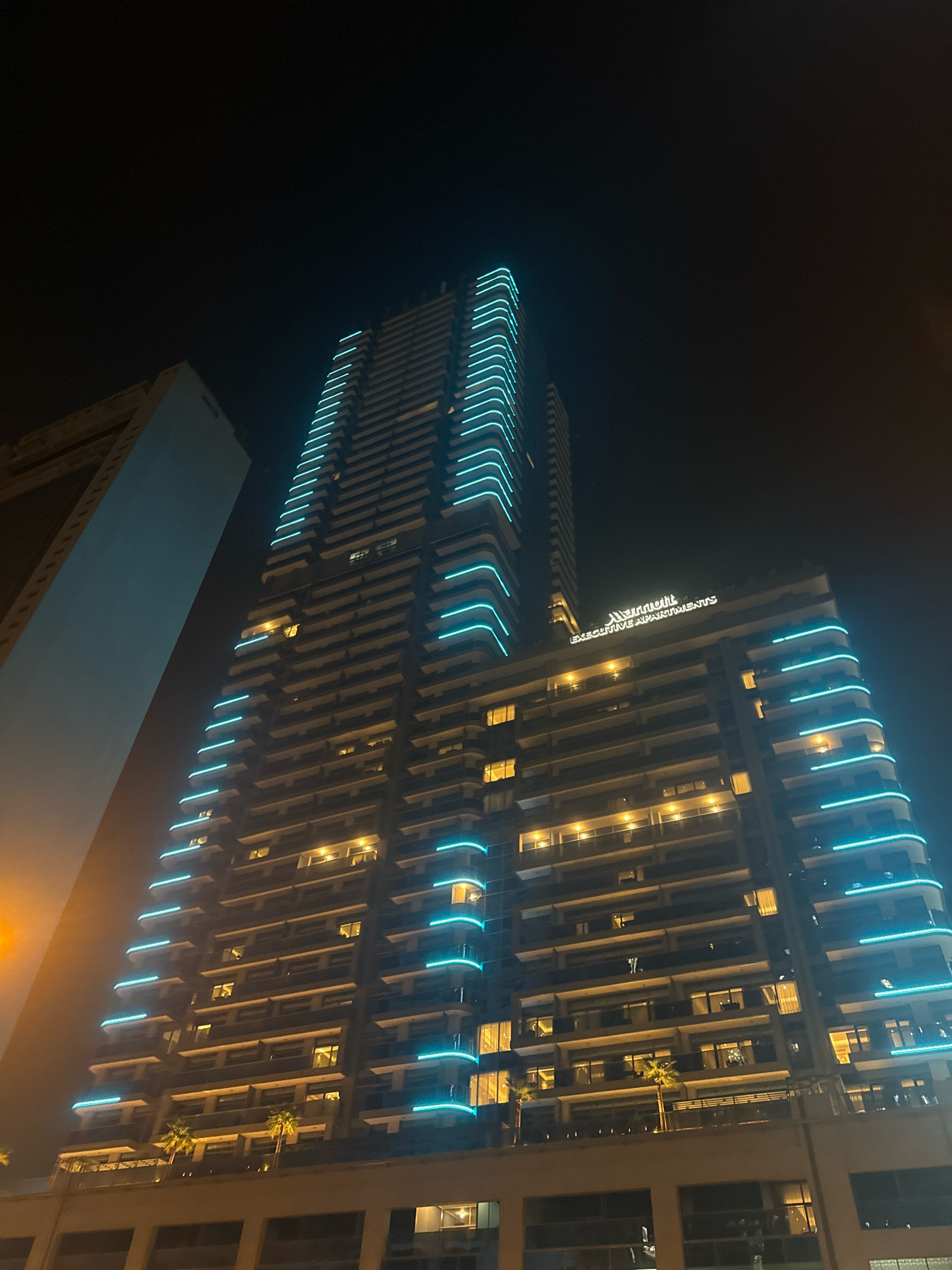 An outdoor, low-angle, night-time shot captures the Marriott Executive Apartments building in Dubai, United Arab Emirates. The building is illuminated by blue neon lights outlining its balconies and by yellow lights from within its many windows. The facade of the building features a stepped design with a prominent row of balconies on each level. To the left of the Marriott building, a portion of another tall, light-colored building is visible, also at an upward angle. The sky is dark, typical of a nighttime scene.
