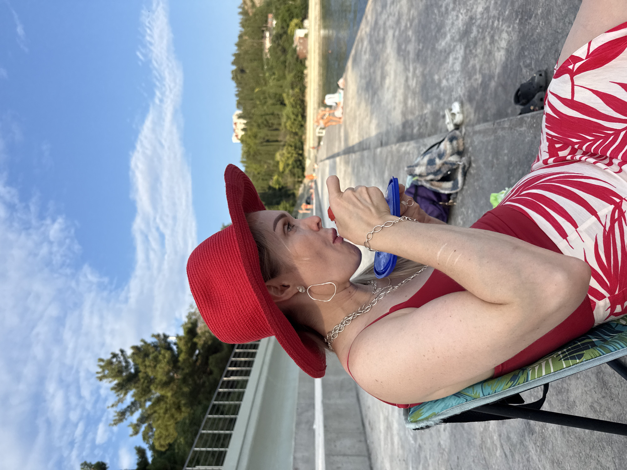 An adult female wearing a red hat, red top, and a red and white patterned skirt is seated in a folding chair. She is holding a small blue container and appears to be eating from it. She is wearing large hoop earrings and several necklaces. The background shows a concrete walkway next to a body of water. In the distance, there are several people sunbathing and trees on a hillside with buildings. The scene is outdoors, under a blue sky with scattered clouds. The location is identified as Utios, Ukraine.