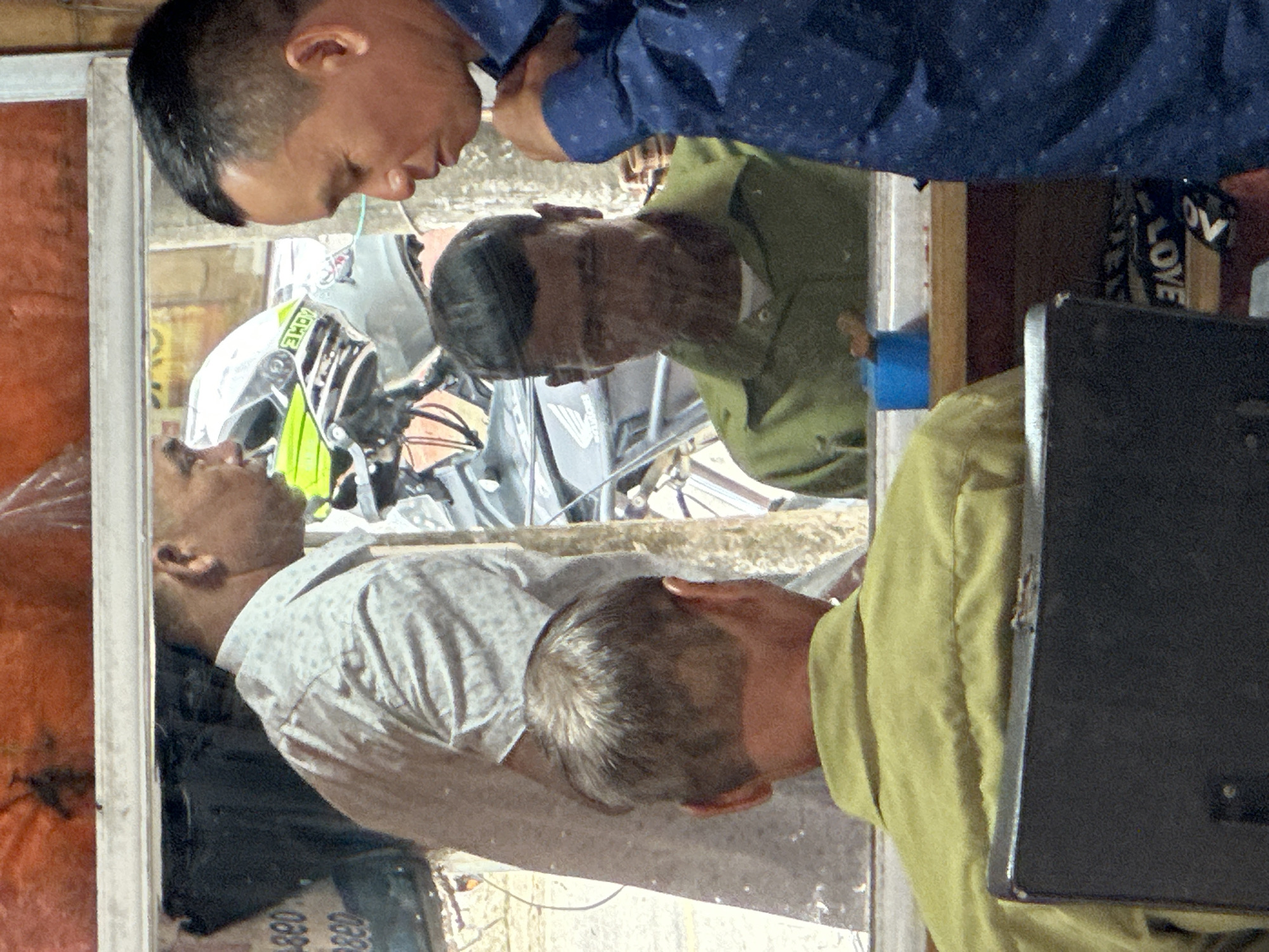 The image, taken in Bắc Yên, Vietnam, shows three adult males positioned around a glass window or mirror. In the foreground, a man with short graying hair faces away from the camera, wearing a lime green collared shirt and seated behind a black object, possibly a chair or counter. The reflection in the window reveals another man with short dark hair, also wearing a lime green collared shirt, looking downwards. To the right of this reflection, a man with short dark hair and a blue patterned collared shirt is visible from the side, looking down and to his right. Behind the man in the green shirt's reflection, a man with short dark hair and a light-colored patterned collared shirt is seen in profile, looking to the left. A motorcycle helmet with yellow and green accents is also visible in the reflection, positioned behind the man in the light-colored shirt. The background appears to be an indoor setting, with some items partially obscured by reflections.