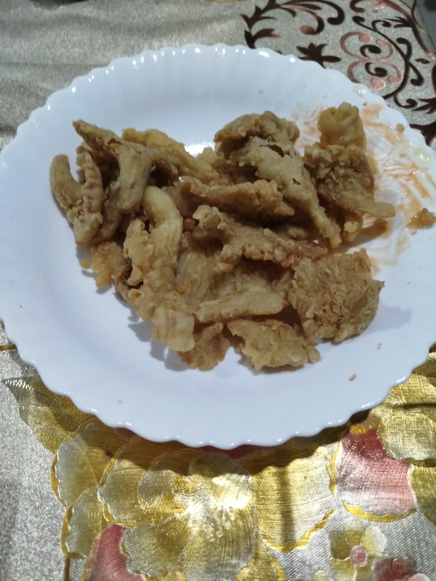 A white plate filled with fried mushrooms sits on a table covered with a gold and red floral-patterned tablecloth. The mushrooms are golden brown and appear crispy. Some reddish sauce is visible on the plate, likely a dipping sauce or residue from serving. The background is blurred, but suggests a casual dining setting. The image does not provide enough information to determine its location in Bandungrejo, Indonesia.