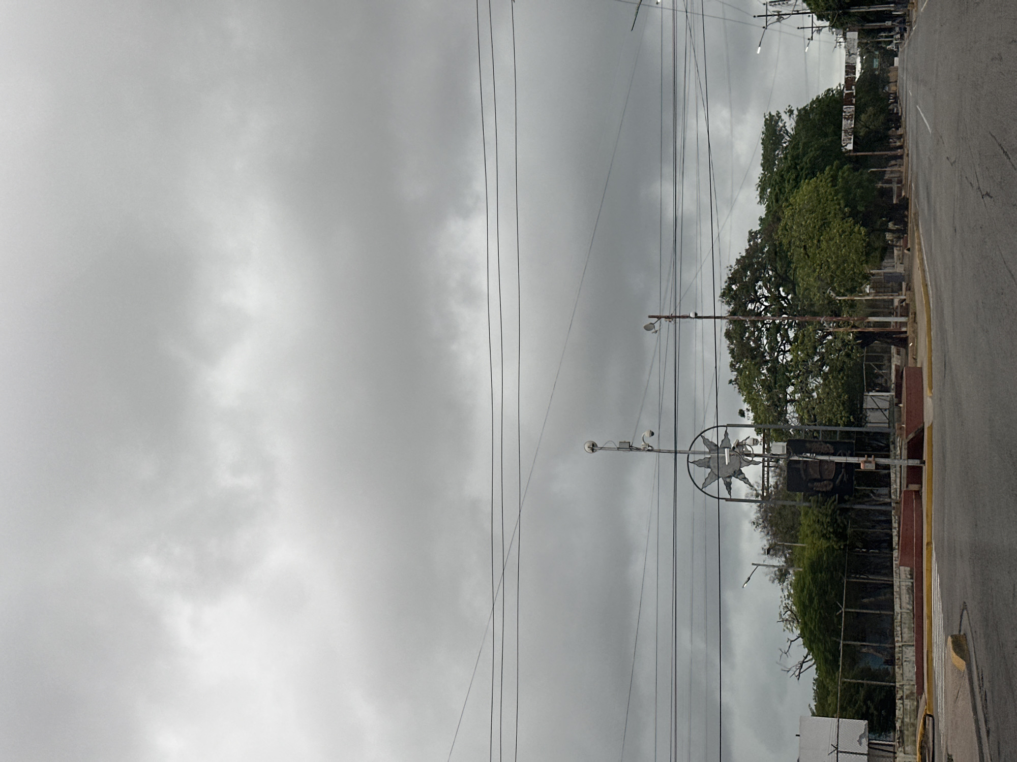 The image shows a street scene in Juan Griego, Venezuela, under a heavily overcast sky. In the foreground, a paved road curves to the right, with yellow and white road markings and a pedestrian crossing. To the left, a concrete barrier separates the road from a fenced-off area containing lush green trees and some vegetation. A white building with a fence is visible behind the trees.
Dominating the center of the frame is a monument with a distinctive star-shaped metal sculpture at its apex. The monument is surrounded by trees and appears to be located in a public space. Multiple utility poles with wires stretch across the sky, and streetlights are visible. The overall atmosphere is somber due to the cloudy weather. No people are clearly visible in the image.