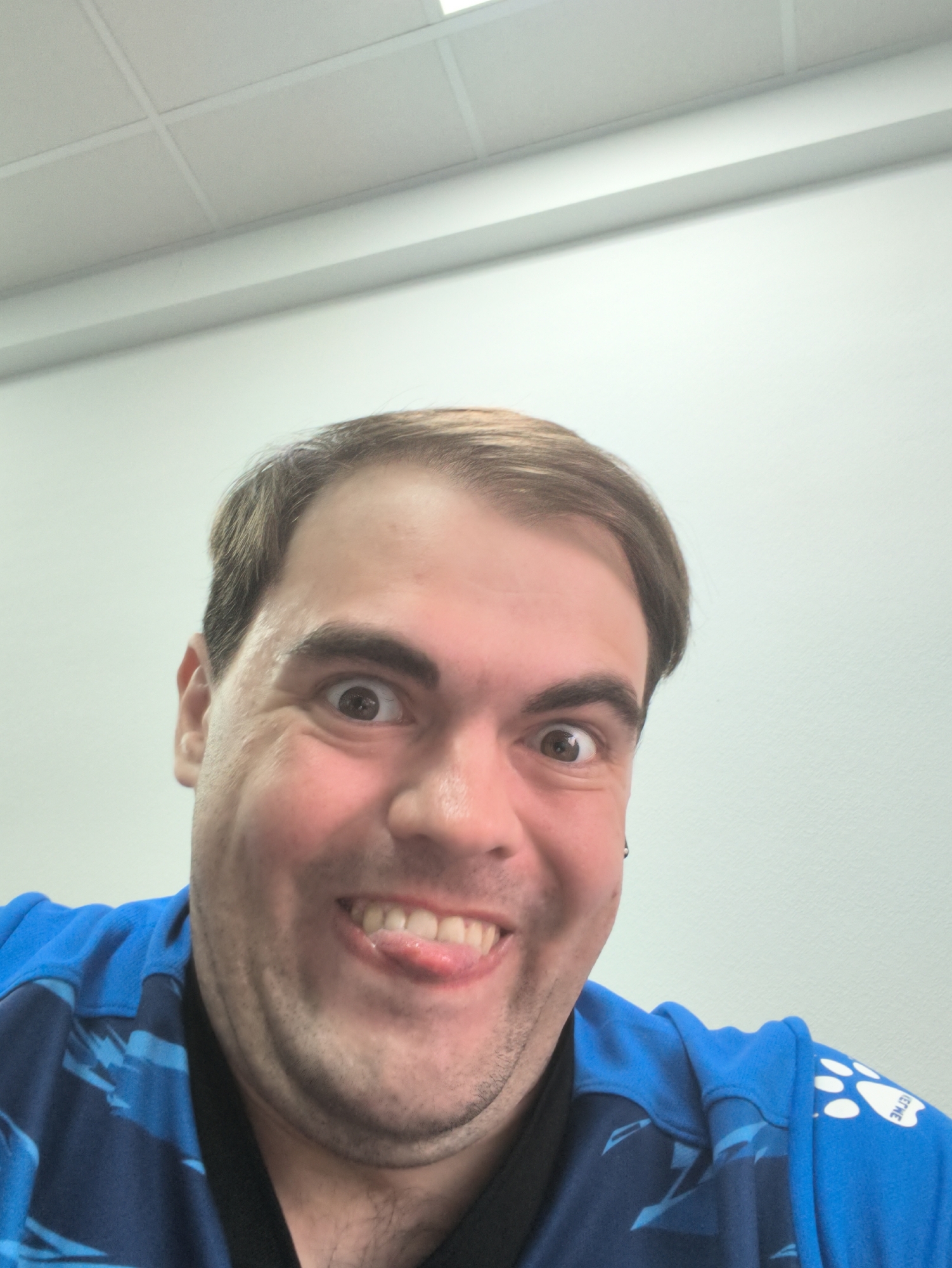 A close-up, front-facing selfie of a man with fair skin and brown hair is shown. He is wearing a blue shirt with a dark blue pattern and a white paw print logo on the shoulder. The man is making a goofy face, sticking his tongue out slightly and widening his eyes. His expression appears playful and lighthearted. The background is a plain, light-colored wall and a ceiling with fluorescent lights, suggesting an indoor setting, possibly an office or a room. The lighting is bright and even, illuminating the man's face clearly. There are no other people or objects of significance in the frame. The image is taken from a slightly low angle, looking up at the man.