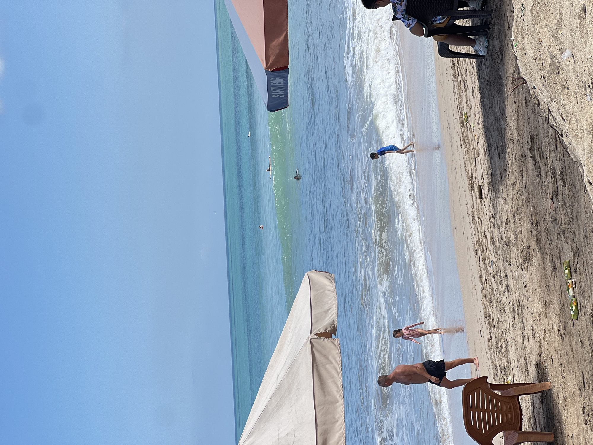 A wide shot captures a sunny day at Kuta Beach, Indonesia. In the foreground, a man and a young girl walk along the wet sand at the edge of the turquoise ocean. The man, shirtless and wearing black shorts, walks towards the left of the frame. The girl, in a long-sleeved pink swimsuit, walks slightly ahead of him, heading towards the water. A child in a blue swimsuit is visible further down the beach, near the waves. Two large beach umbrellas, one beige and one with brown and blue panels, provide shade. The umbrella with brown and blue panels has the words "SANTI BAR" printed on its side. Several people are in the ocean, with some surfing on small waves. The water is clear and blue, with white surf. The sand is a light brown color, with some dried leaves and small offerings scattered on it in the foreground. In the right foreground, a person is seated in a dark chair, facing away from the camera. The sky is a clear, bright blue.