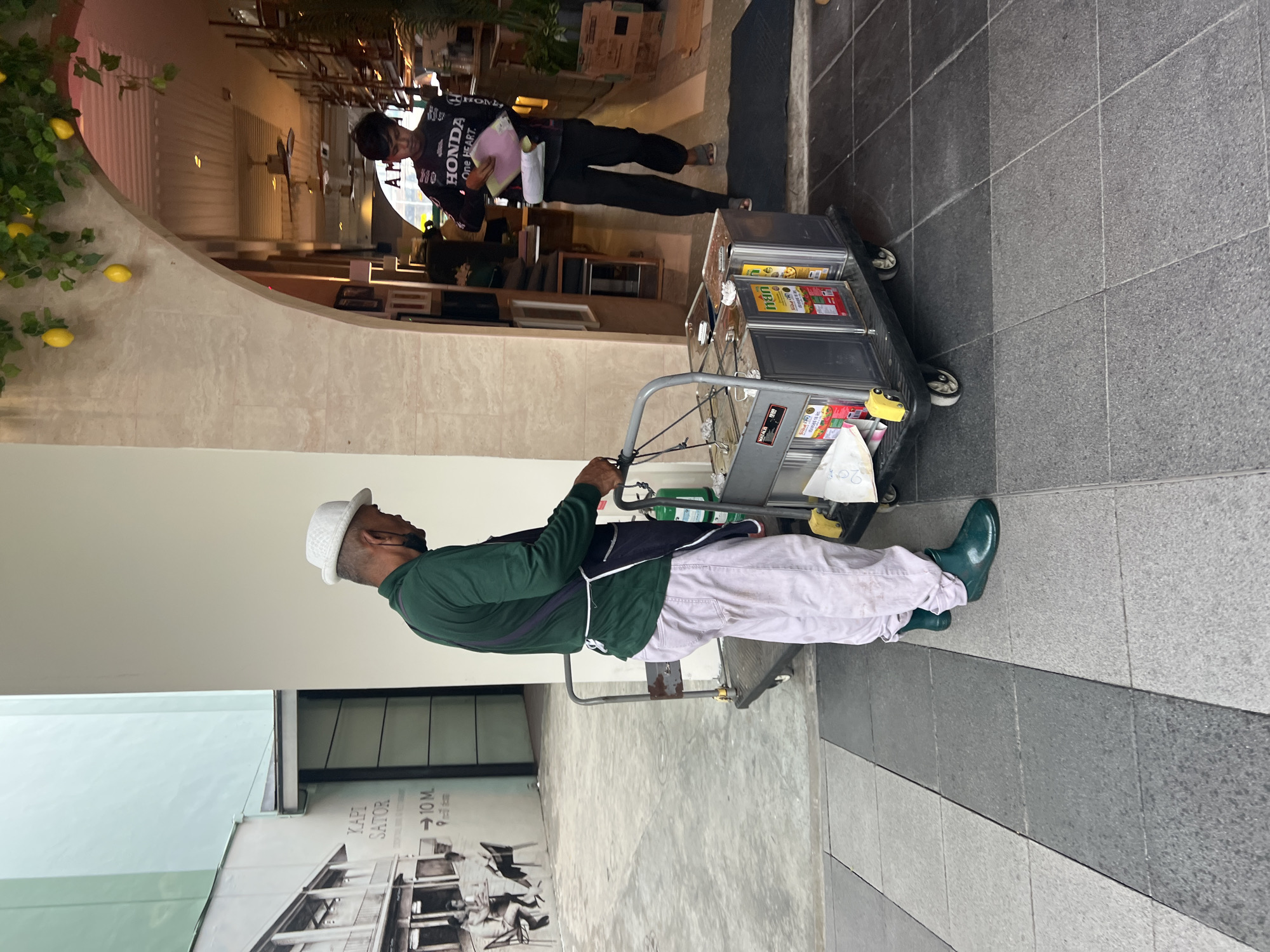 An adult male wearing a white fedora hat, green long-sleeved shirt, light-colored pants, and green boots stands to the left of a two-tiered metal cart filled with various items, including what appear to be large bottles of cooking oil. He is facing the cart and appears to be pushing it. To the right of the cart and further into the background, another adult male wearing a black long-sleeved shirt with "HONDA" prominently displayed and black pants is walking into an establishment with an arched entrance. The establishment appears to be a restaurant or shop with a lit interior and shelves. The scene takes place on a paved walkway outside, with a light green wall and a black and white mural visible on the left side of the frame. Lemons hang from a decorative vine above the arched entrance. The overall environment suggests an outdoor commercial area in a tropical location, possibly Ko Samui District, Thailand.
