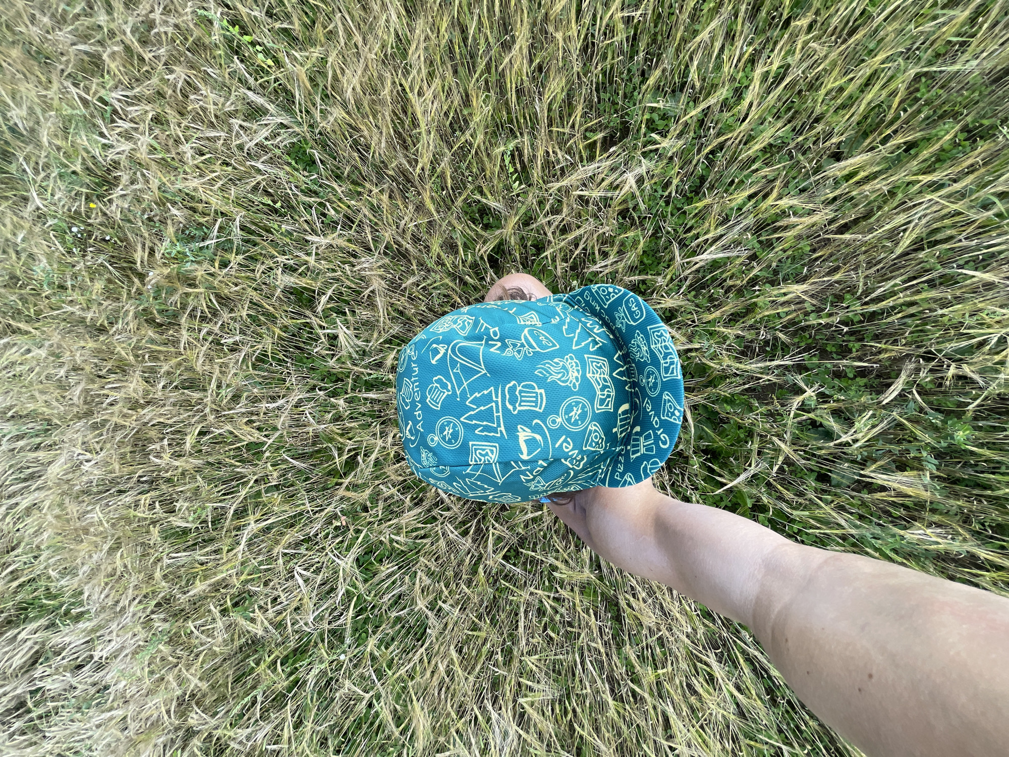 In Maksimovo, Russia, a person's arm is visible in the foreground, holding a teal baseball cap adorned with white illustrations of adventure-themed items such as compasses, tents, coffee cups, and beer mugs. The cap is held over a field of tall, dry grass. The background consists entirely of this grassy field, with some green foliage interspersed. The image is taken from a high-angle perspective, looking down.