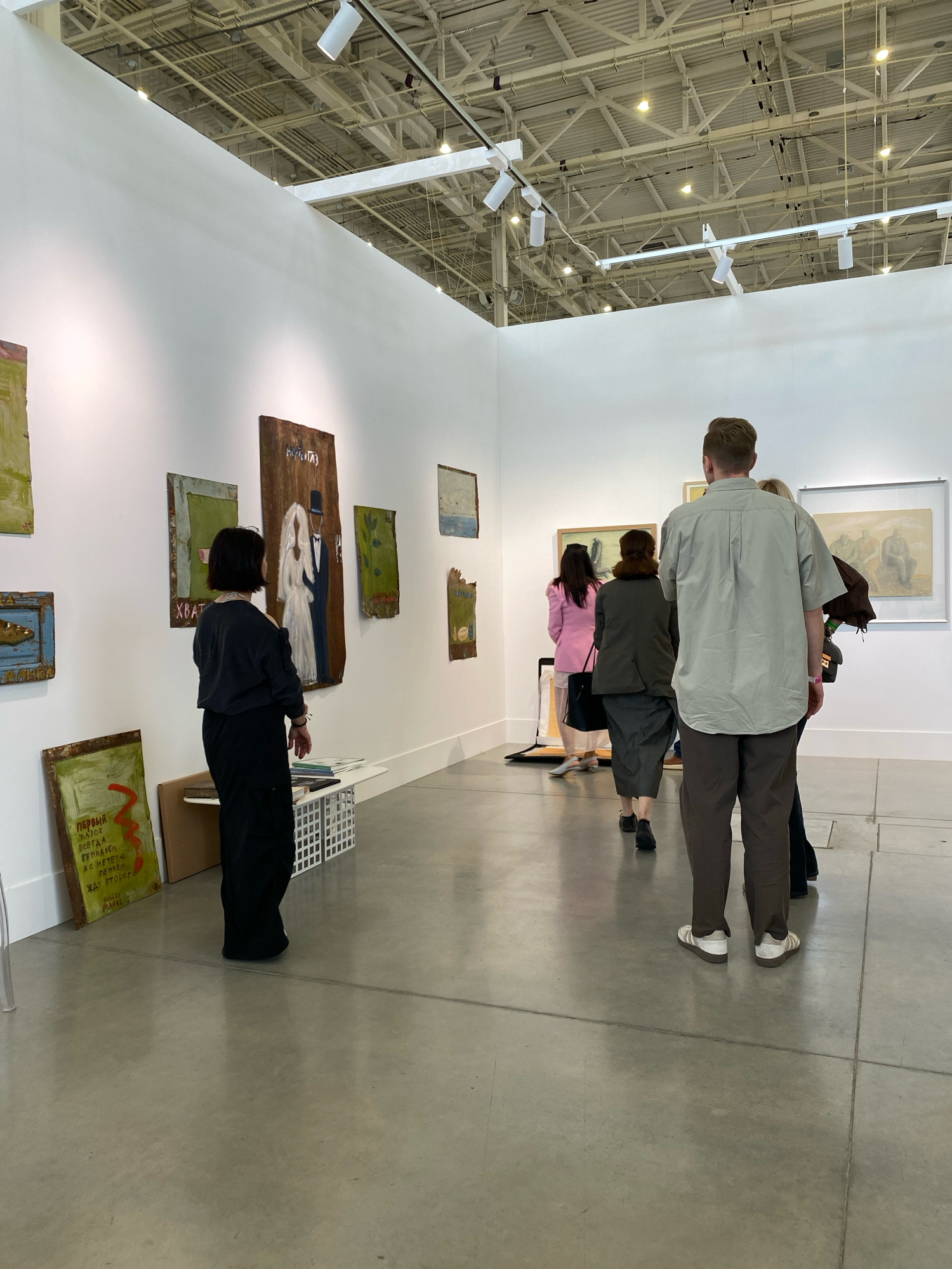 In an art gallery in Khimki, Russia, several individuals are observing artwork displayed on white walls. The space features a polished concrete floor and an industrial ceiling with exposed ductwork and track lighting.
In the foreground, a man wearing a light olive-green short-sleeved button-up shirt and dark brown trousers stands with his back to the camera, facing the artwork. He is wearing white sandals. To his left and slightly behind him, another person, partially obscured, appears to be standing.
Further into the gallery, on the left side of the image, a woman with short black hair wearing a dark top and black trousers stands facing a painting depicting a bride and groom. To her left are several other artworks, including one with text in Cyrillic. A white slatted storage unit is positioned near her feet.
On the right side of the gallery, a group of people are viewed from behind. A woman in a pink jacket, another in a dark grey jacket and a long grey skirt, and a third individual are looking at paintings on the wall.
The artwork itself consists of various paintings and possibly mixed-media pieces, many on distressed or textured surfaces, with some featuring figurative elements and text. The overall impression is of a contemporary art exhibition.