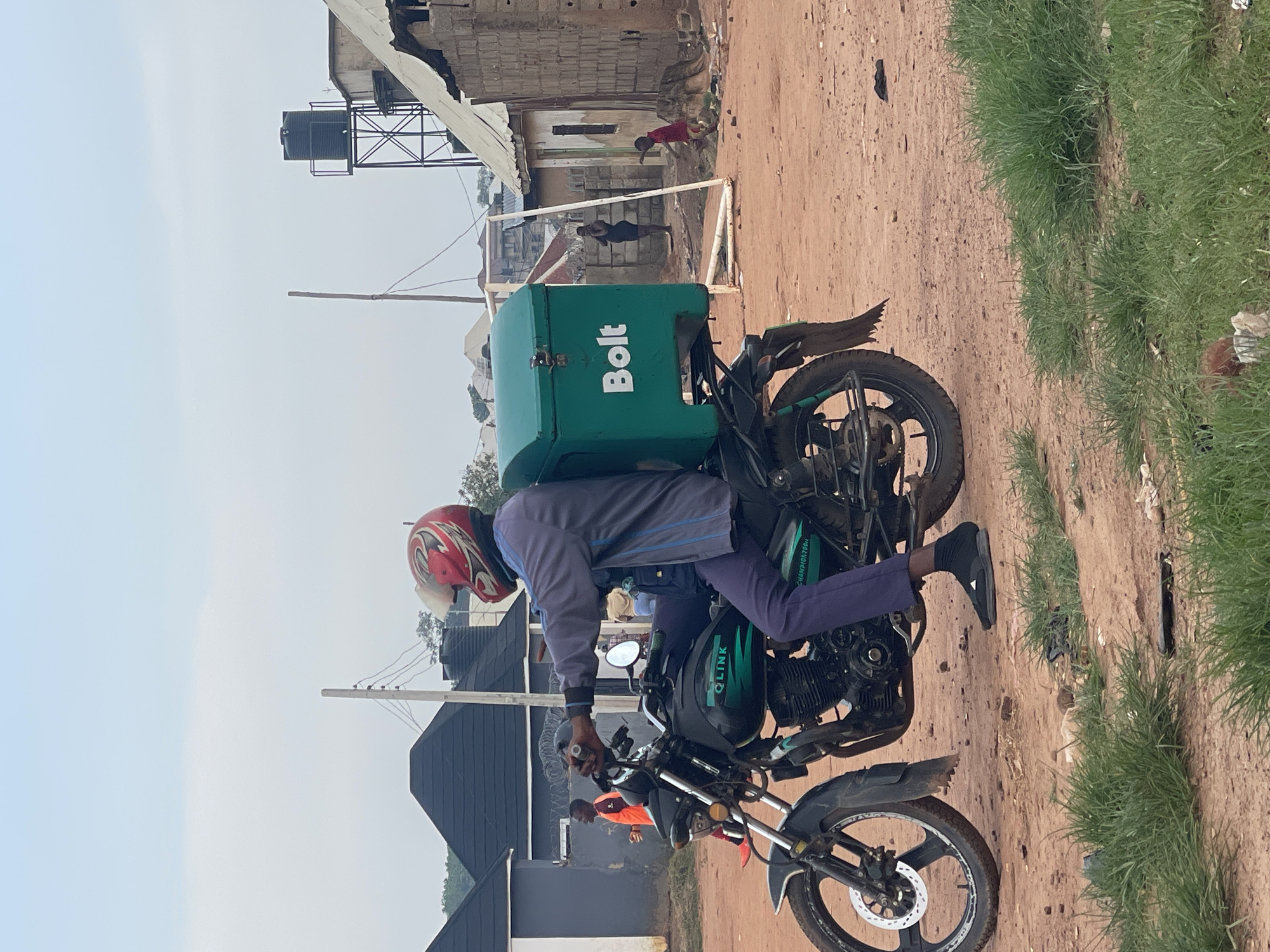 A Bolt delivery person is riding a motorcycle with a green delivery box on the back. The motorcycle is black with green lettering that says "Qlink". The delivery person is wearing a gray jacket, blue pants, black shoes, and a red and white helmet. They are on a dirt road in a residential area. In the background, there are several houses, a water tower, and some people walking around. The sky is overcast. This scene is located in Kurunduma, Nigeria.