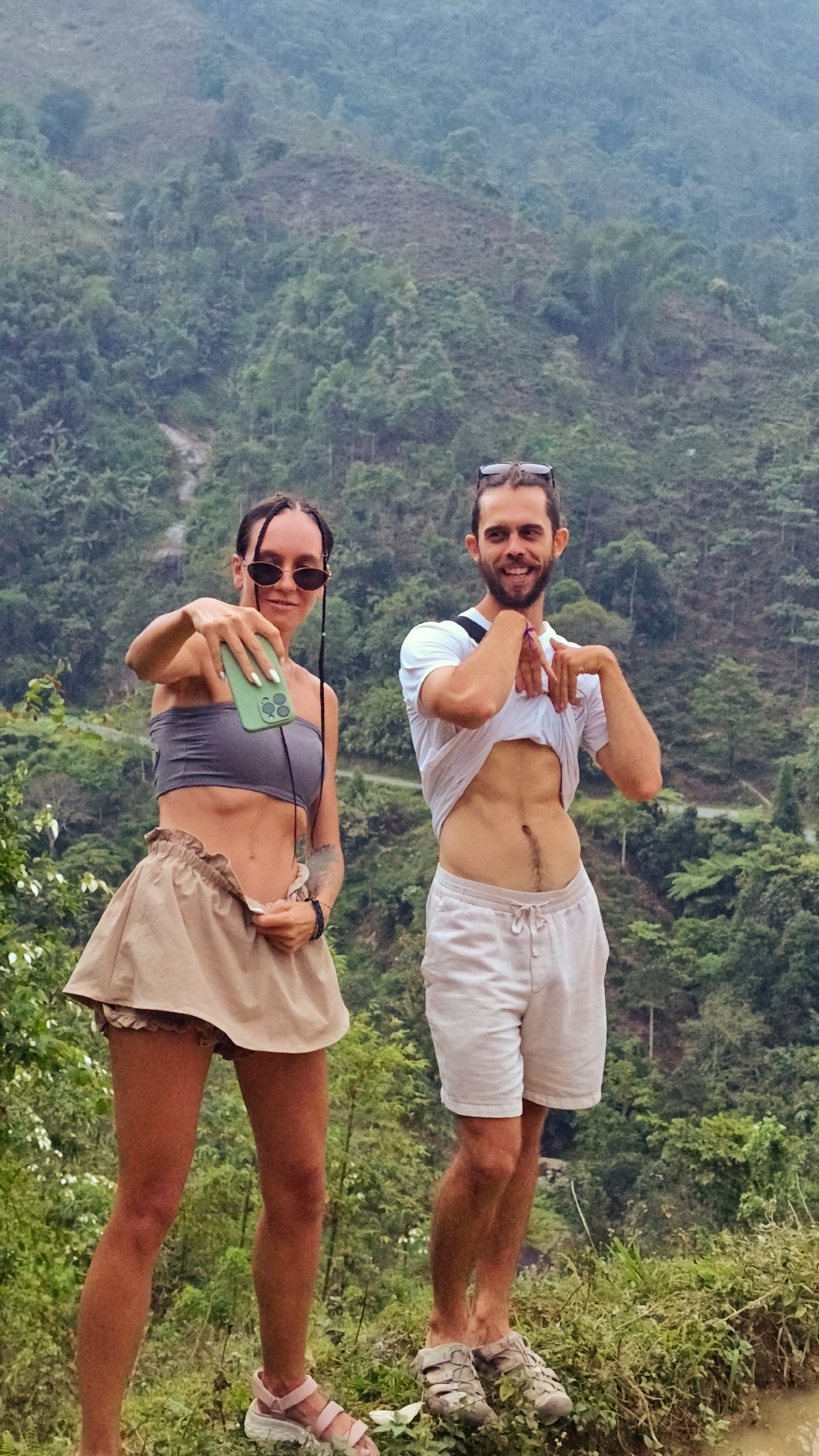 A full-length outdoor portrait captures two smiling individuals, a woman and a man, posing playfully against a backdrop of lush green mountains in Nậm Dịch Commune, Vietnam. The hazy, overcast conditions cast a soft, natural light on the scene.
On the left, a woman with long dark hair, wearing sunglasses, a grey bandeau crop top, and a light brown ruffled skirt, holds a bright green smartphone forward as if taking a selfie or video. She has a tattoo visible on her left arm and wears light pink chunky sandals. Her expression is a joyful smile.
On the right, a bearded man with sunglasses pushed onto his head wears a white t-shirt and light-colored shorts. He has pulled his t-shirt up to expose his midriff and is making a humorous hand gesture with his hands cupped in front of his chest, grinning broadly. He wears light-colored sandals.
The background reveals a dense, verdant mountainous landscape, with layered hills covered in tropical foliage, characteristic of the region. A winding dirt path or road is faintly visible among the distant trees. In the immediate foreground, there is natural vegetation and a patch of muddy ground, possibly part of a rice paddy or water feature. Both subjects exude a relaxed and happy mood, clearly enjoying their outdoor adventure. No visible text is present in the image.