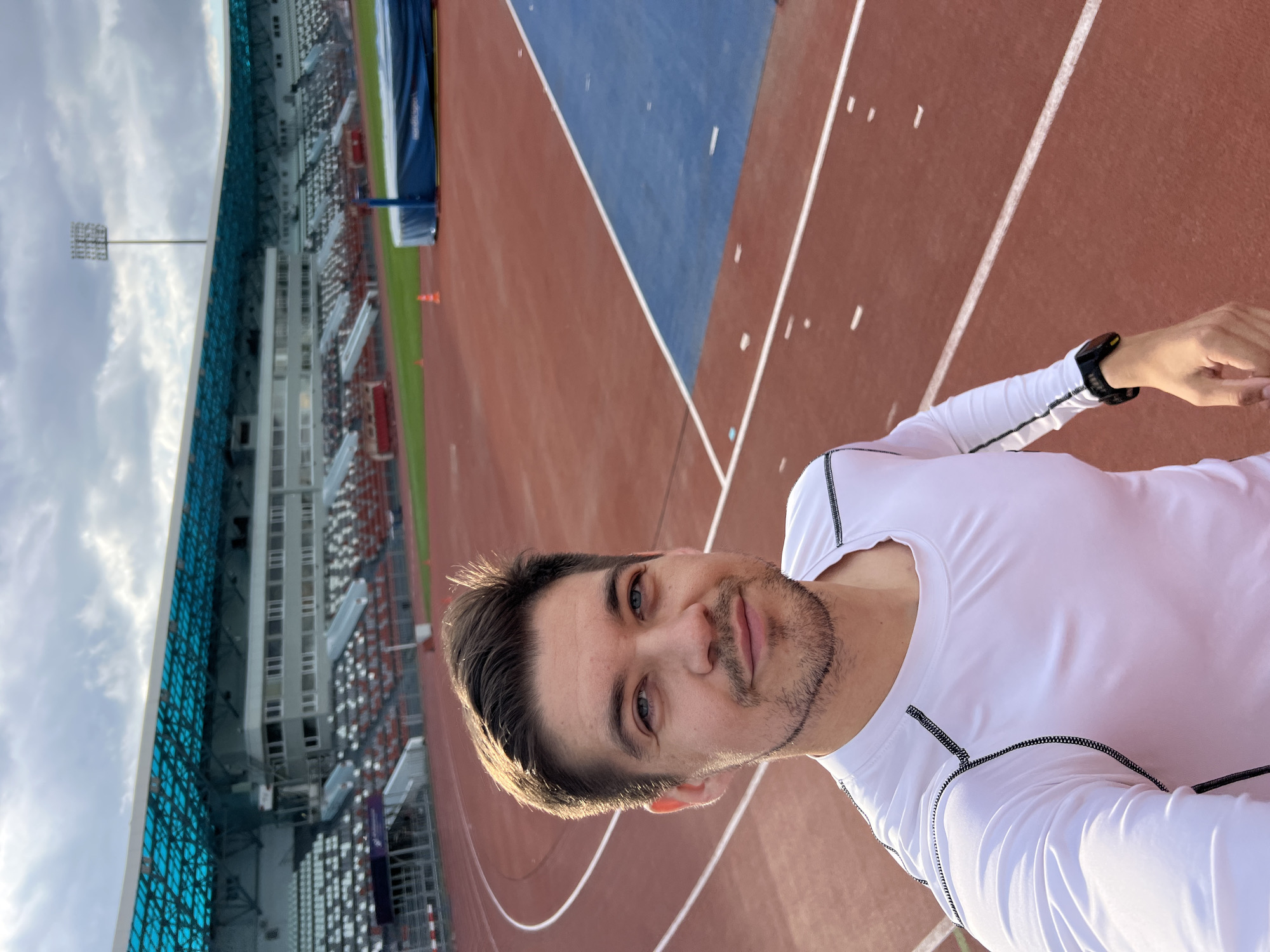 The image is a selfie taken by a fair-skinned man with brown hair and a beard, wearing a white long-sleeved shirt and a smartwatch on his left wrist. He appears to be standing on a red running track at an athletic stadium. The track has white lane markers. A blue area is visible alongside the track, possibly for jumping events. In the background, there are stadium seats filled with rows of red, white, and grey chairs and a metal facade covered in light blue tiles. A stadium light pole is positioned behind the seating. The sky is overcast. The location is likely within the city of Kazan, Russia.