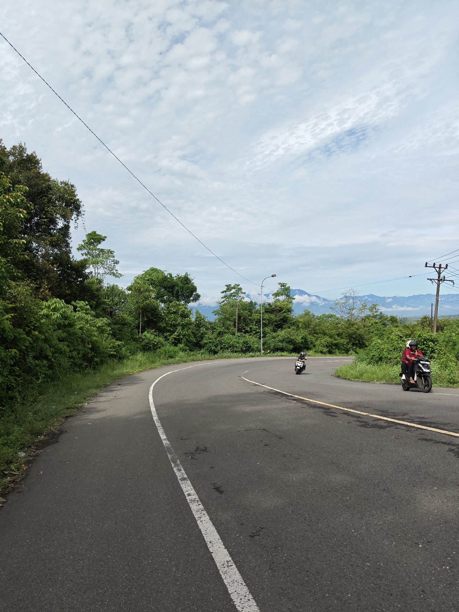 The image depicts a paved road winding through a lush, green landscape in Lamtamot, Indonesia. The sky is partly cloudy. There are two motorcycles on the road. One has two riders wearing helmets, with the front rider wearing a red shirt, further down the road. The other has a single rider and is further down the road. There are trees and dense vegetation on either side of the road. Distant mountains are visible in the background. Power lines run overhead. A road lamp is visible in the middle ground. A white line marks the edge of the road on the left side, and a yellow line divides the road.