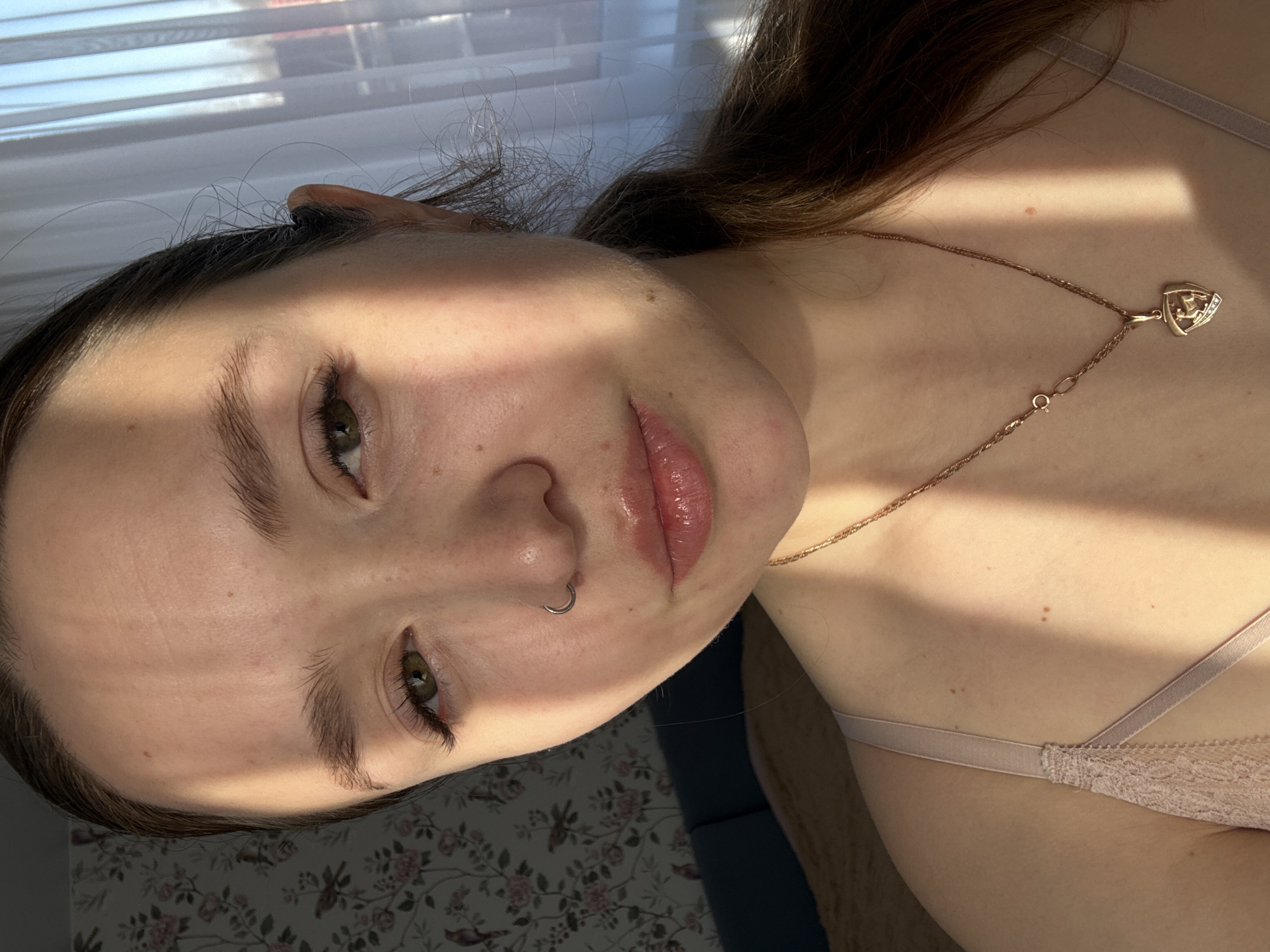 A young woman is featured in a close-up shot. She has light skin, brown hair, and green eyes. She wears a nose ring and a necklace with a pendant. The lighting suggests she is near a window with blinds. The background includes a floral pattern and a hint of blue. No explicit location details pertaining to Izhevsk, Russia, can be identified from the image.