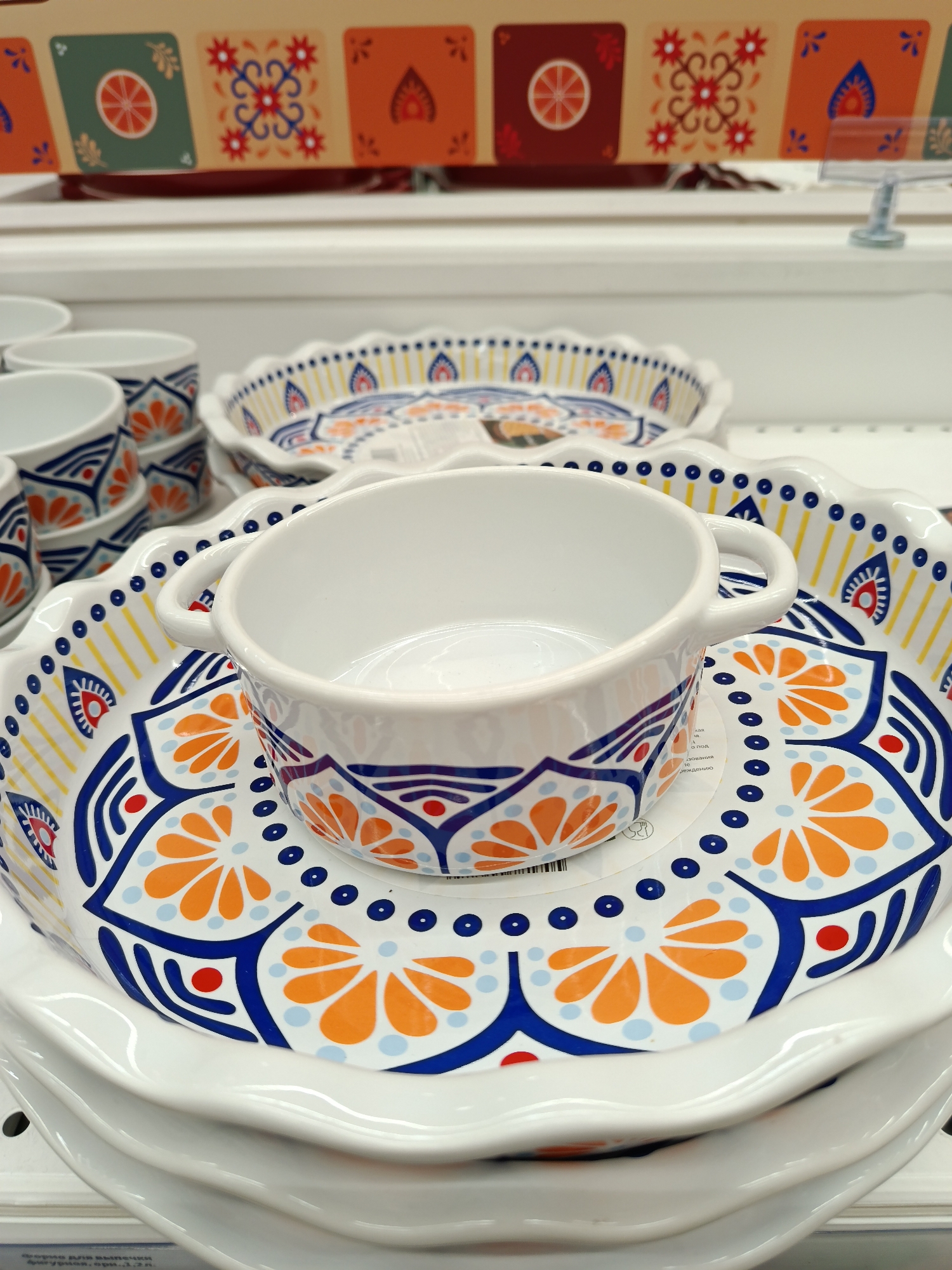 The image depicts a stack of white ceramic plates and a matching bowl, adorned with an orange and blue floral pattern. The items are displayed on a shelf, likely in a retail setting. A box with similar patterned images sits in the background. Based on text visible on a tag in the lower left corner, the scene is located in Moscow, Russia.