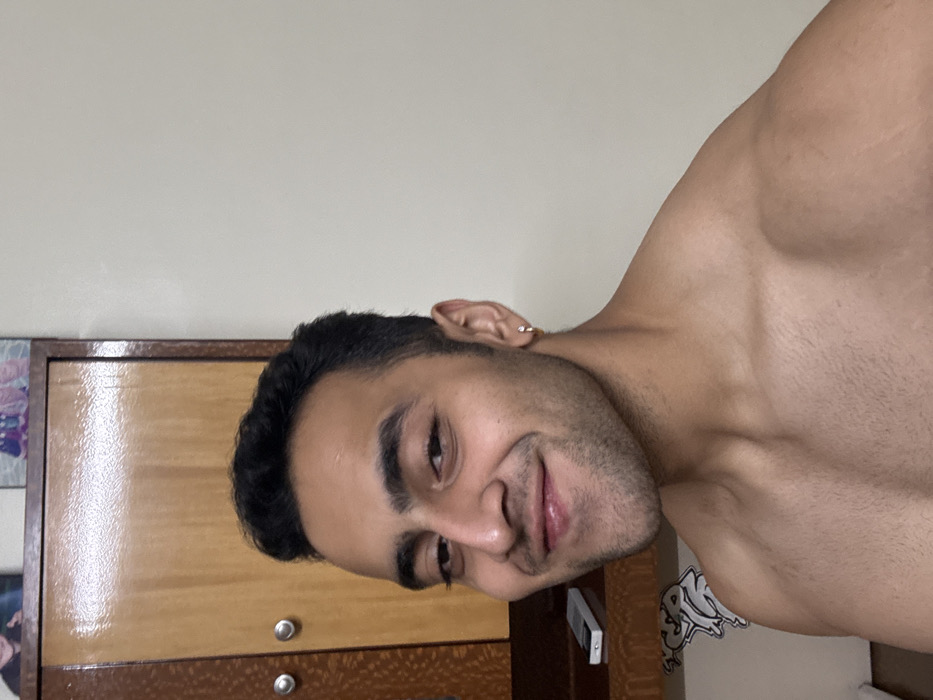 A young adult male is captured in a close-up, rotated image, shirtless and smiling faintly towards the camera. He has dark, short hair, prominent eyebrows, and light stubble on his face. A small silver hoop earring is visible in his left ear. His well-defined shoulders and chest indicate a muscular physique.
The setting appears to be indoors, likely a personal room. A plain light beige or off-white wall forms the background. To the left of the man, a wooden cabinet or wardrobe is partially visible, featuring two lighter-toned wooden doors above two darker-toned drawers, all equipped with silver circular handles. Below his chin, a small white rectangular object, possibly a remote control or phone, rests on a surface. On the right side, behind his shoulder, a white sticker with partially visible black graffiti-style text, possibly reading "SPN," is affixed to a surface. The overall lighting is soft and even, suggesting indoor conditions. The man's expression conveys a sense of confidence and a slight smirk.