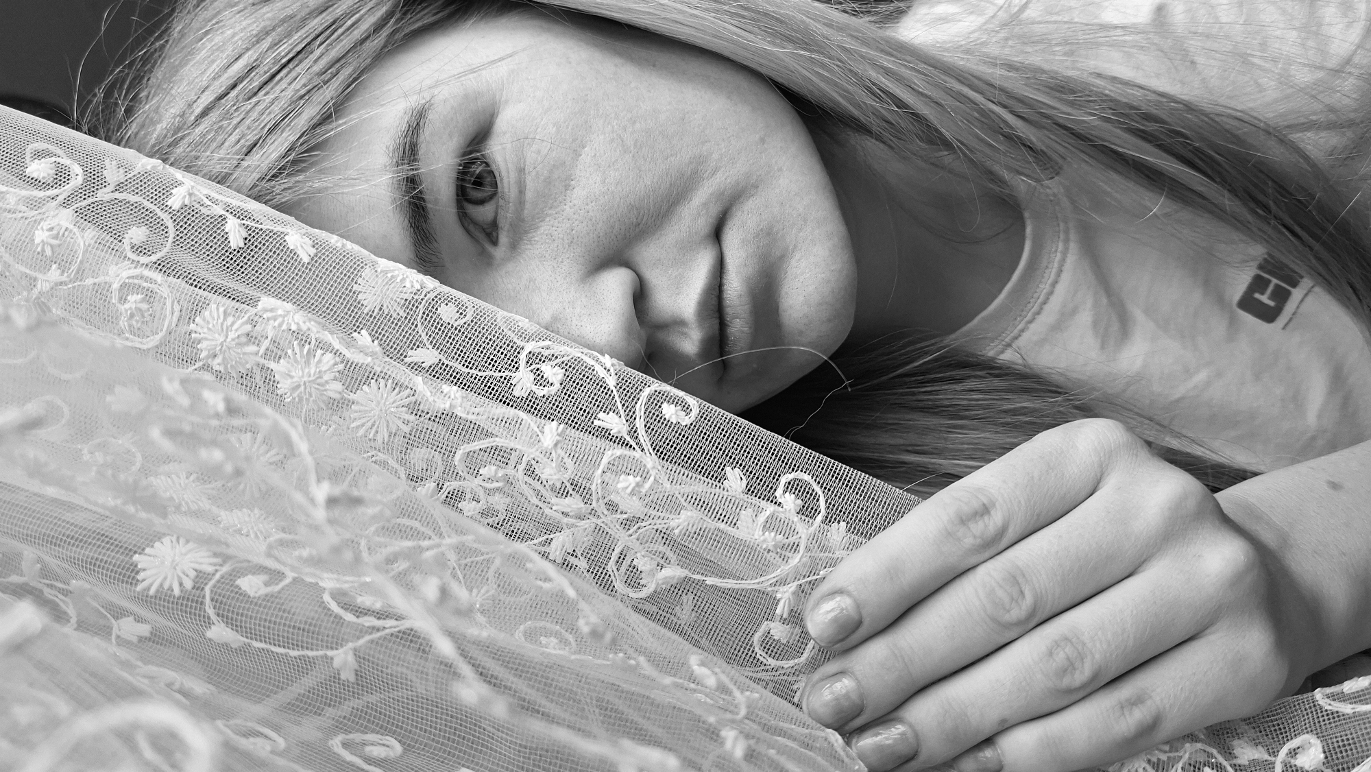 A grayscale close-up depicts a woman lying with her face partially covered by lace fabric. Her left eye is visible, and she is wearing a t-shirt with a symbol on the right side of the collar. Her right hand rests on the lace. The location in the city of Большой Букор, Russia, cannot be determined from the image.