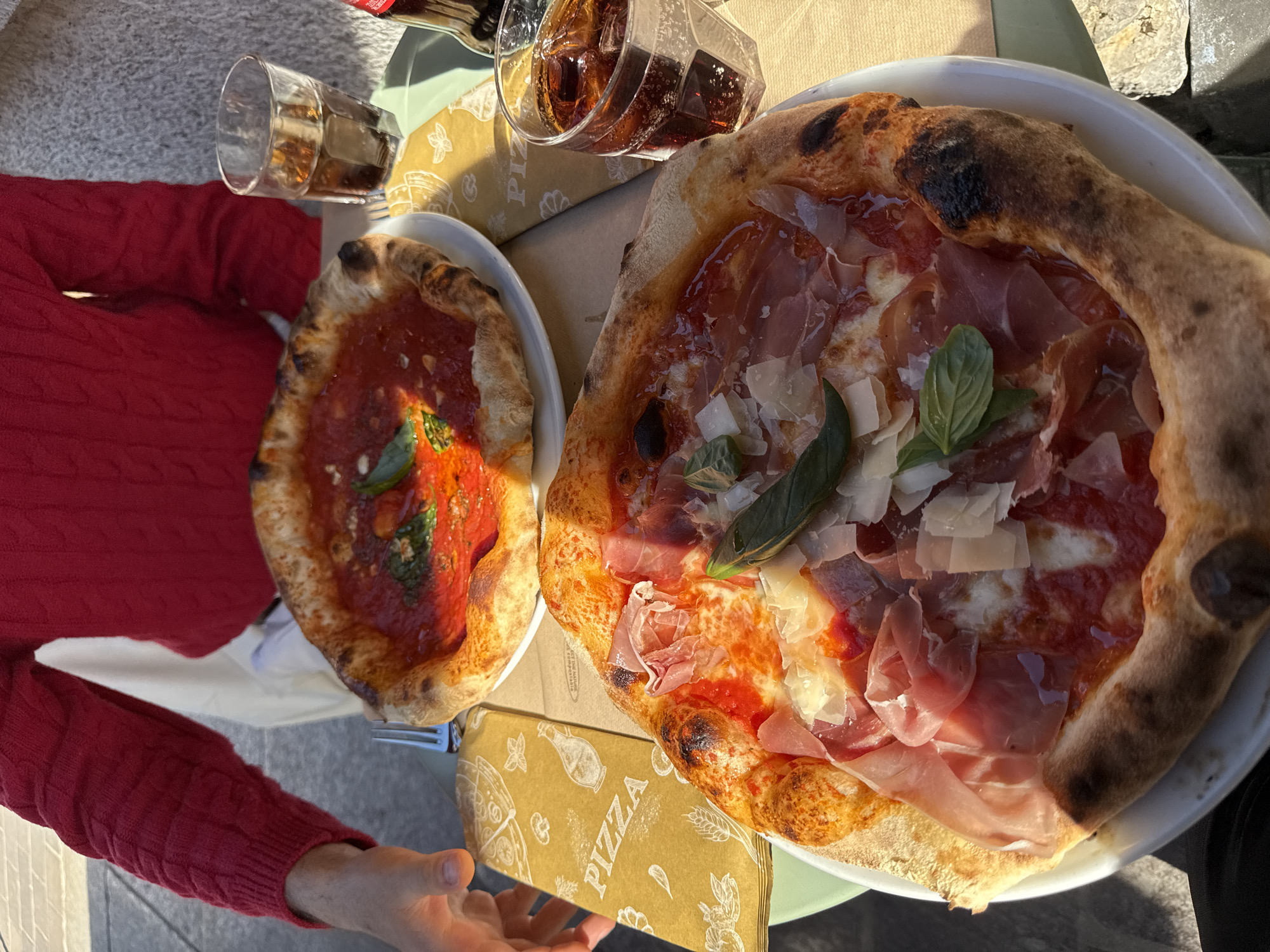 The image captures an overhead view of an outdoor dining scene in Milan, Italy, featuring two freshly baked pizzas on a light green table under bright sunlight.
In the foreground and to the right, a large, generously topped Neapolitan-style pizza rests on a white plate. It boasts a puffy, charred crust, a vibrant red tomato base, melted mozzarella, numerous slices of prosciutto ham, and flakes of shaved Parmesan cheese, garnished with a few fresh basil leaves. The sunlight creates a glossy sheen on its surface.
To the left, a smaller pizza, also on a white plate, appears to be a simpler Margherita. It features a rich tomato sauce and a couple of fresh basil leaves on its surface, with a similarly thick and slightly charred crust.
A person is partially visible in the frame, wearing a red long-sleeved, cable-knit sweater. Their arms and hands are seen near the bottom and left side of the table, suggesting they are seated and about to eat or are in the process of dining.
Two glasses containing a dark, carbonated beverage with ice cubes are present. One glass is nearly full, while the other is approximately half-empty. A small bottle, likely containing the same beverage, is also on the table.
Underneath the pizzas, a light brown paper placemat or box is visible. It features various line-art illustrations of food items and the word "PIZZA" printed in a decorative font across its surface. The strong shadows cast by the objects indicate that the photograph was taken on a clear, sunny day, likely in the afternoon. The scene conveys a casual and enjoyable outdoor meal.