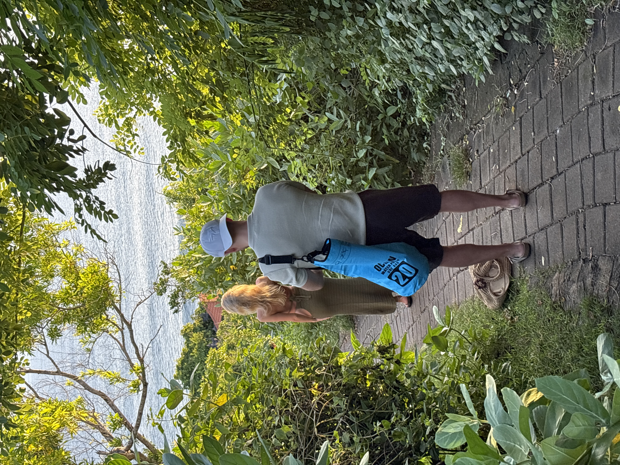 The image shows a man and a woman standing on a paved path lined with dense green foliage, with a view of the ocean visible through the trees. The man is wearing a light-colored long-sleeved shirt, dark shorts, a white baseball cap, and sandals. He is carrying a blue bag labeled "Water Proof Dry Bag 20" over his shoulder. The woman, standing slightly behind him, is wearing a light olive-green dress. The path is constructed from rectangular gray bricks. The background includes a glimpse of the ocean and some buildings through the vegetation. Given the description of Kuta Selatan, Indonesia, as a tourist destination known for beaches and lush landscapes, it is likely that this image depicts a coastal area in that city.
