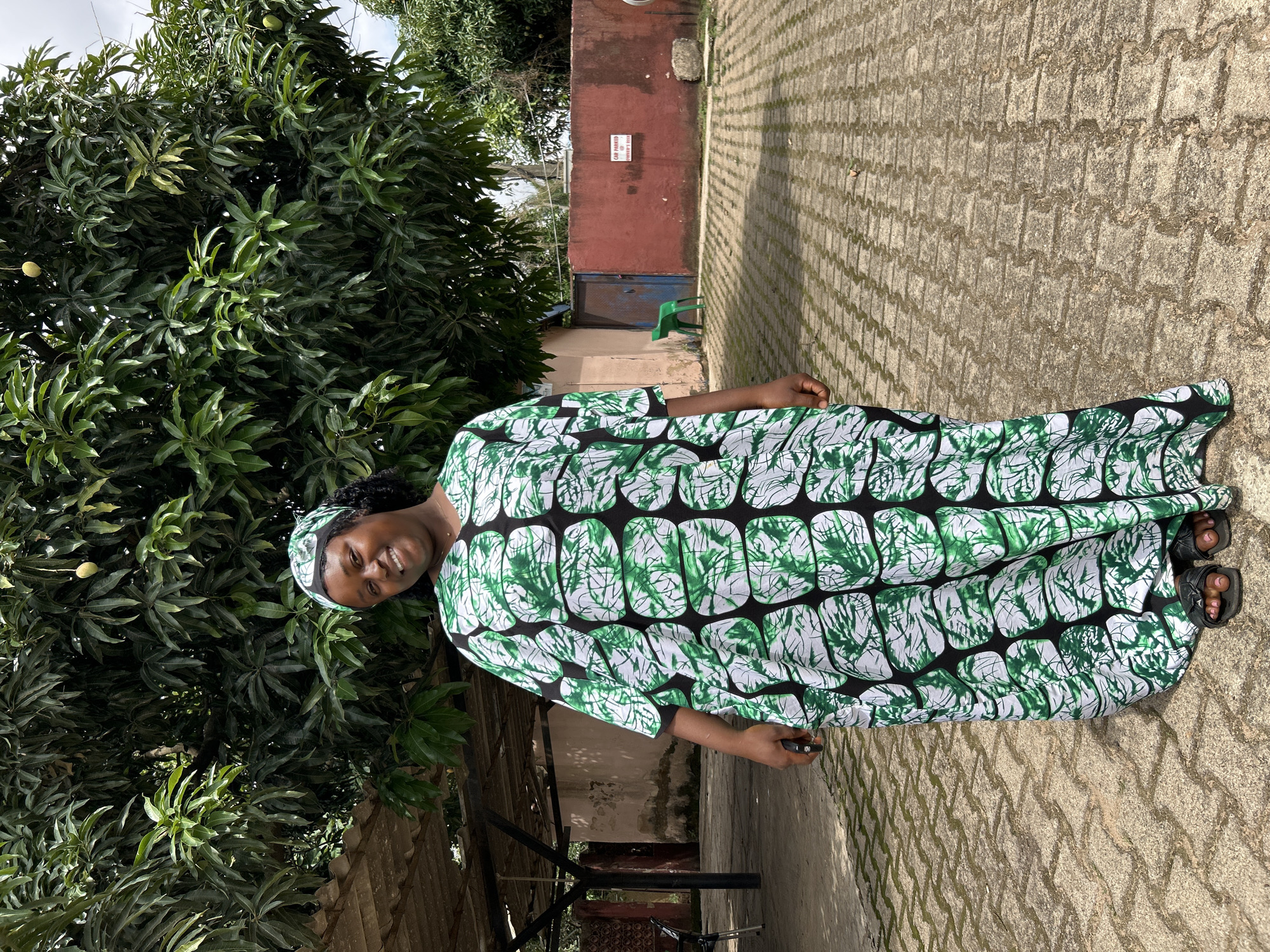 A woman stands centered in the frame, outdoors in Jos, Nigeria, smiling at the camera. She wears a long, loose-fitting kaftan-style dress and a matching headscarf, both patterned with green and white abstract shapes outlined in black. Her left hand holds a small dark object. She wears dark sandals, and her feet are visible.
The ground is paved with interlocking concrete blocks, showing some dirt and vegetation in the crevices. A large, dense mango tree with several unripe mangoes dominates the upper background, primarily on the left. To the far left, a corrugated metal roof supported by dark beams is partially visible, covering an area with a light-colored wall behind it.
In the mid-ground on the right, a red-orange painted wall extends horizontally. A white sign with black text reading "CAR PARKED AT OWNER'S RISK" is affixed to this wall. To the left of the red wall, a closed blue metal gate is visible, with a green plastic chair positioned in front of it. Additional dense foliage is present in the upper right background. The scene is brightly illuminated by natural daylight.
