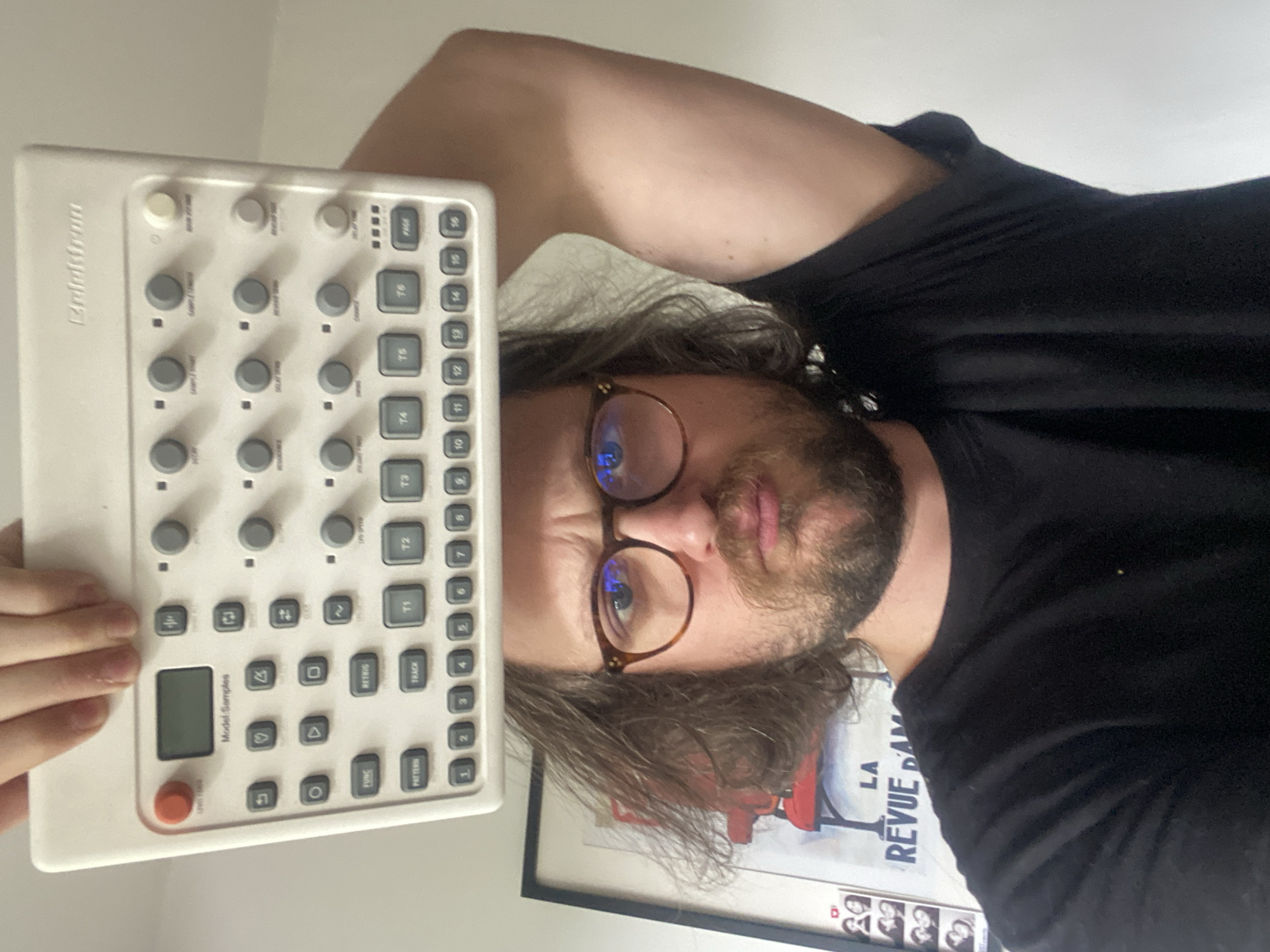 A man with brown hair and a beard, wearing glasses and a black shirt, is holding a white Elektron Model:Samples instrument. A poster with the words "LA REVUE DAM" is visible in the background. The image appears to have been rotated. There is no evidence to suggest that this image was taken in Rusanovo, Russia.