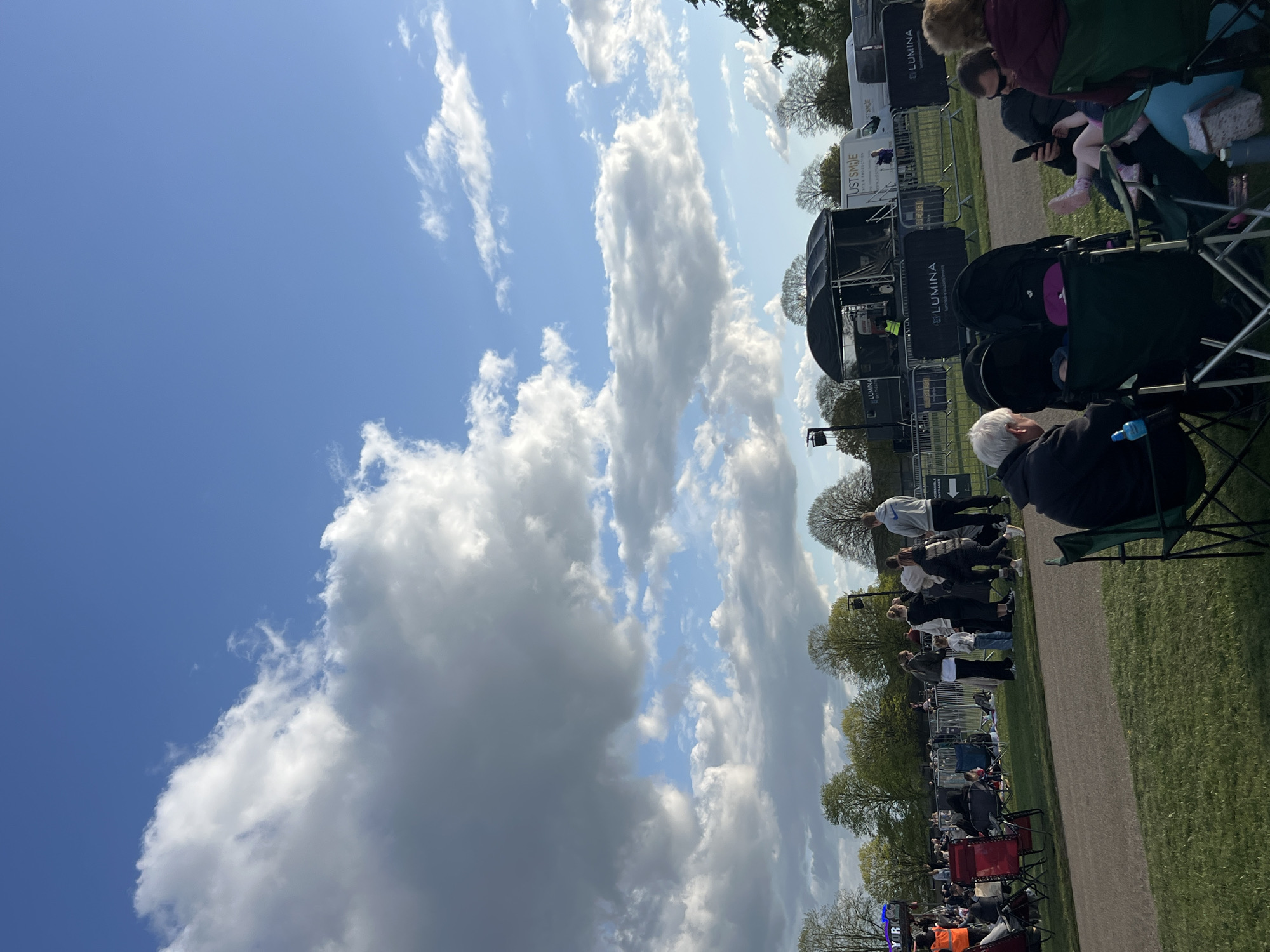 The image depicts an outdoor event taking place in South Norfolk, United Kingdom. A crowd of people are seated in chairs on a grassy area. A stage is set up with sound equipment and signage. The event appears to be a performance or show. The sky is predominantly blue with puffy white clouds. Trees are visible in the background.
