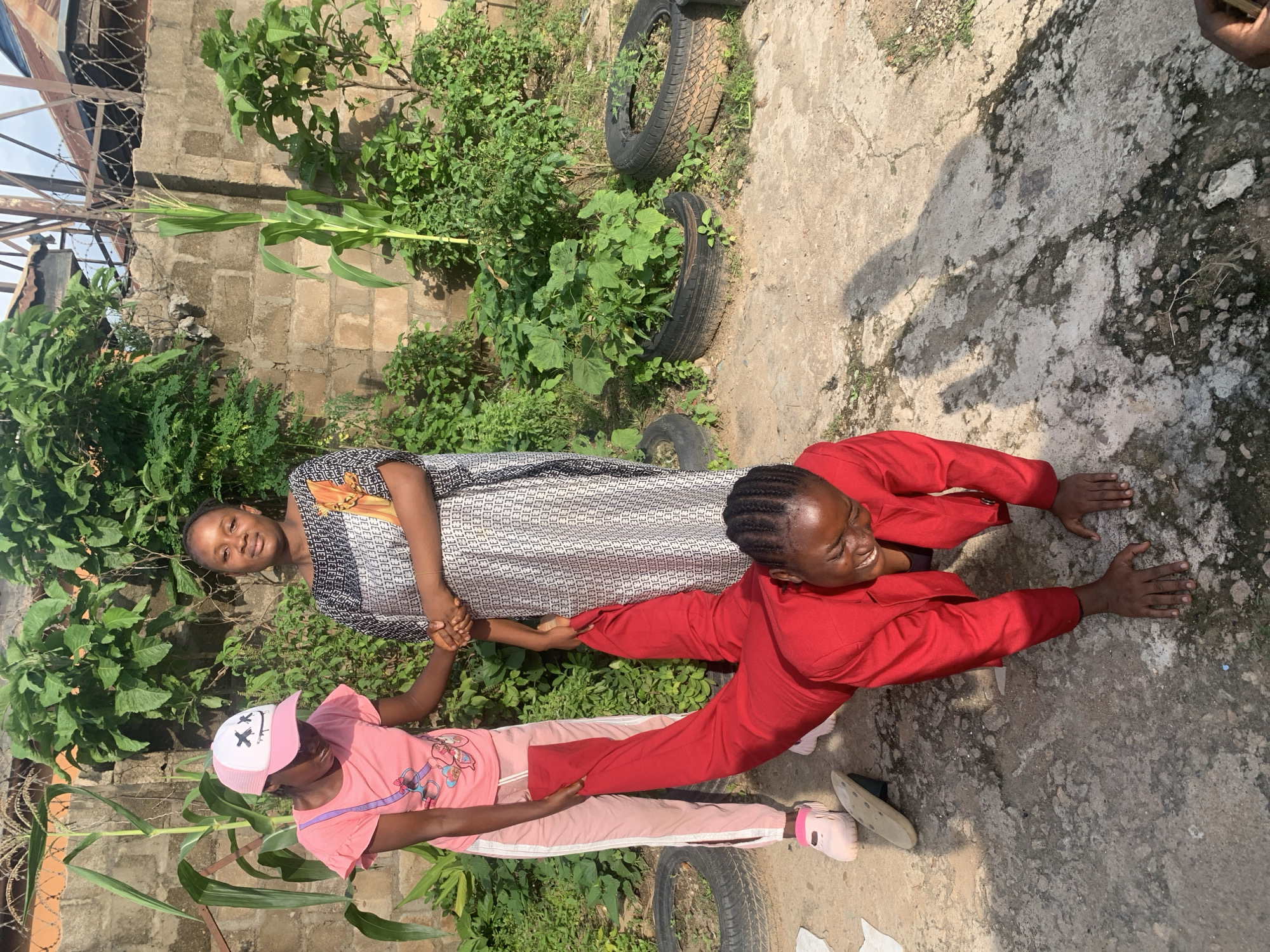 In Sabon Tasha, Nigeria, a young Black woman in a red blazer and pants is on her hands and knees on a concrete surface, laughing. Another young Black woman wearing a pink t-shirt, pink pants with white stripes, and a pink cap with a black smiley face design is standing and holding the first woman's outstretched legs. Behind them, a third Black woman in a patterned dress stands, her arm extended. The setting appears to be an outdoor area with a brick wall and lush green foliage, including a corn stalk, in the background. Several old tires are visible on the ground, with some containing plants. The lighting suggests it is daytime.