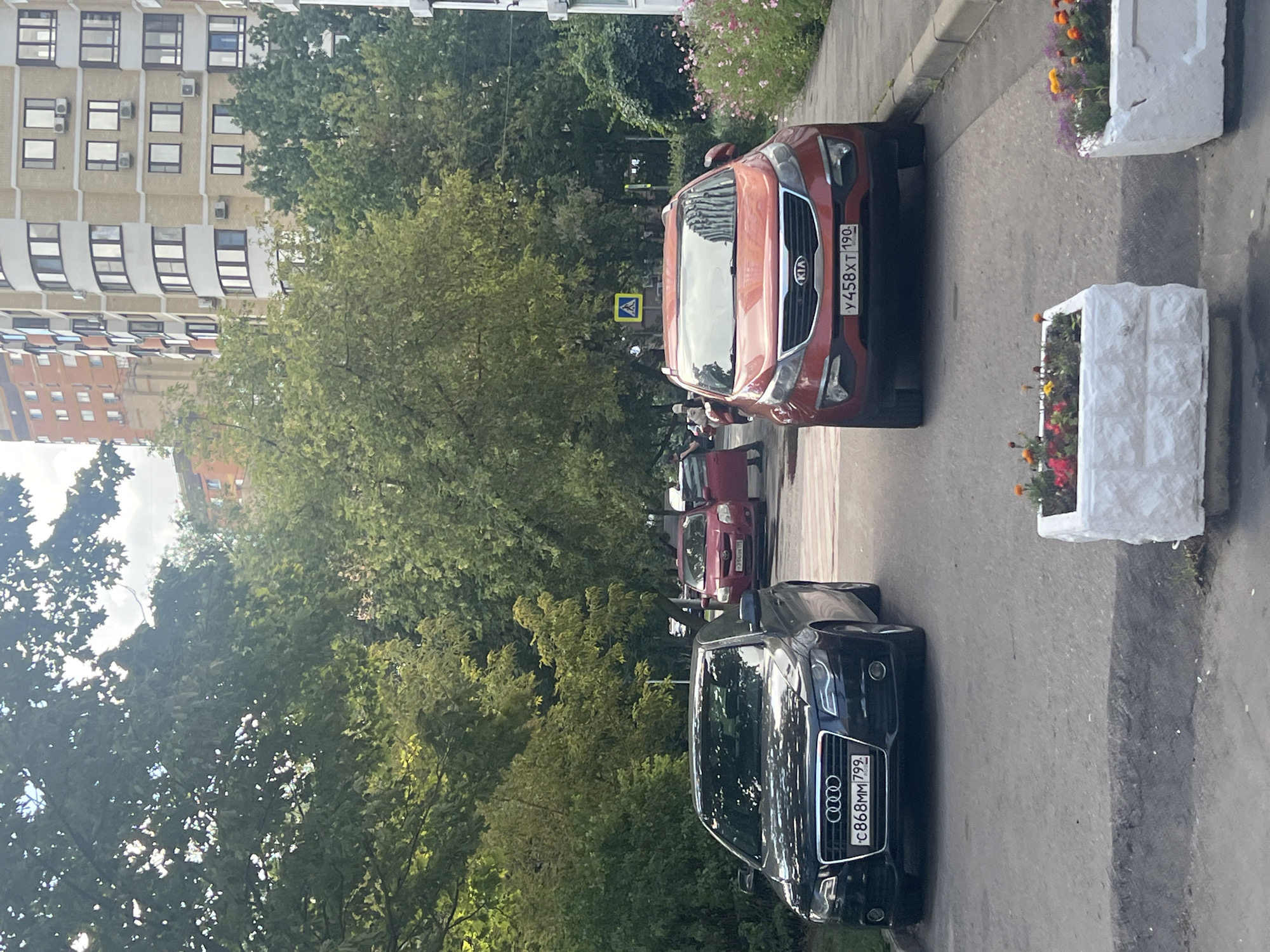 In Moscow, Russia, an outdoor scene depicts a residential area with a multi-story apartment building in the background and lush green trees lining the street. Two cars are parked in the foreground: a black Audi on the left and a red Kia Sportage on the right. The Audi has a license plate that reads "C868MM 799". The Kia's license plate is "Y458XT 190". A red car is visible further down the street, and a pedestrian is standing near it. Two white planters filled with flowers are placed on the pavement in the lower part of the image. The sky is partially cloudy.