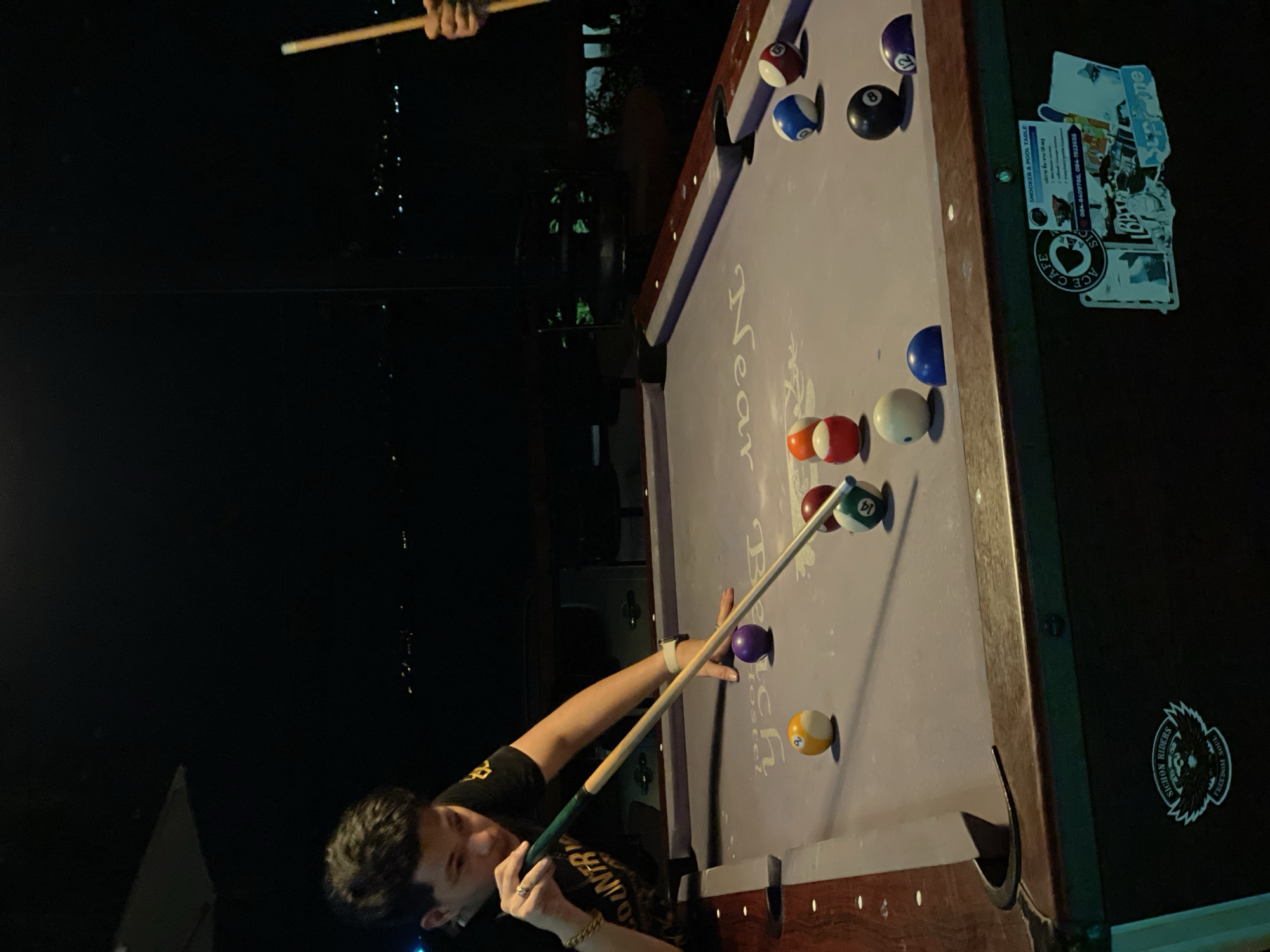 The image depicts a person playing pool outdoors in Baan Bang Rak, Thailand. The pool table is positioned outside, with a dark night sky visible in the background. The pool table is dark wood with a lavender felt. The balls are arranged for a shot, with the cue ball aligned to hit the 14-ball. A person is aiming the cue stick, their arm extended across the table. Stickers are adhered to the side of the table. The OCR identifies text related to the table as "SNOOKER & POOL TABLE," with a phone number, and additional text related to a cafe called "ACE CAFE."