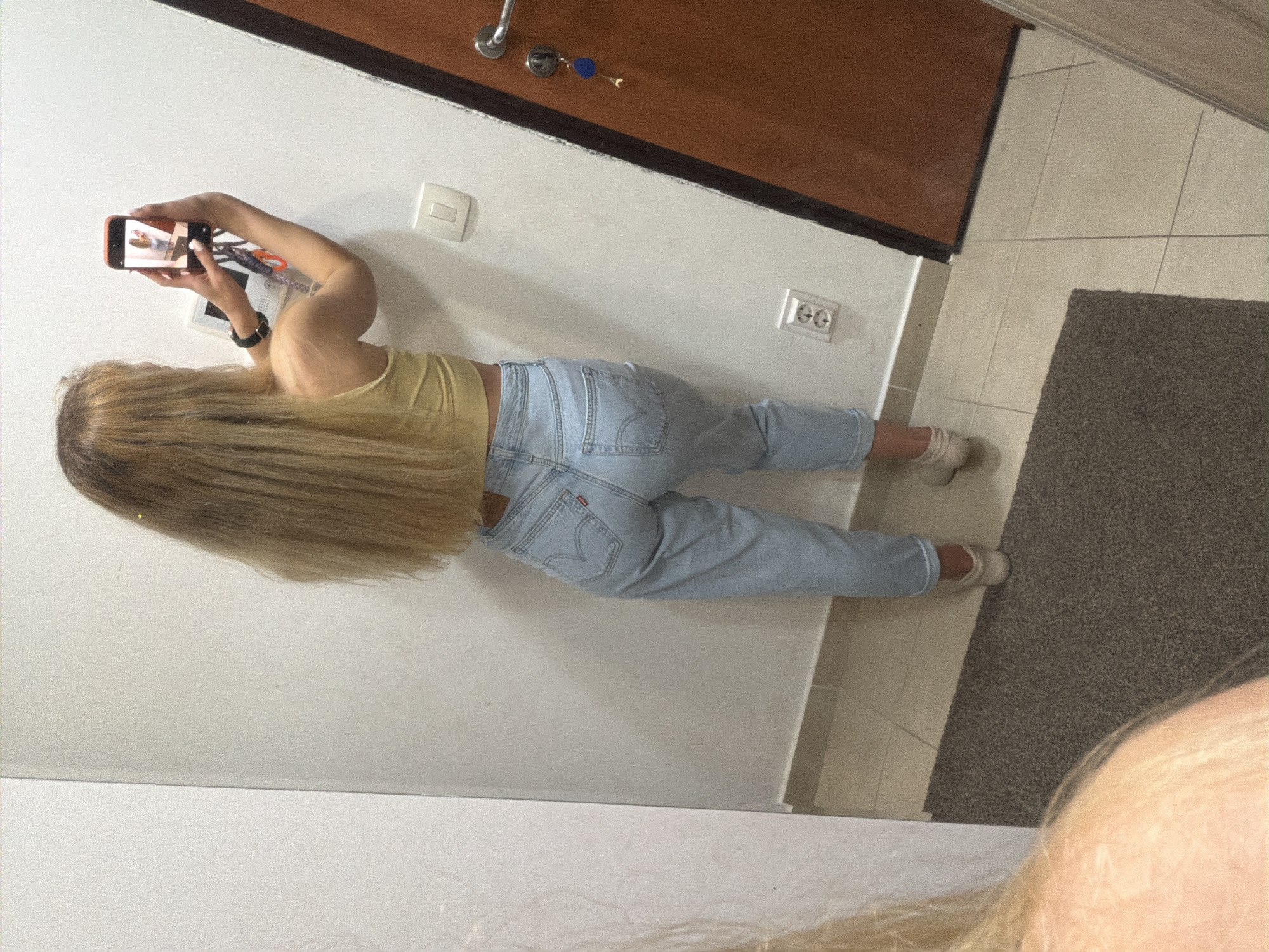 A young woman with long, blond hair is taking a picture of an intercom system on a white wall using a smartphone. She is wearing a yellow top, light blue jeans, and white socks. The wall is located near a door with a brown frame, white light switch, and a European-style power outlet. In the upper left corner, keys are visible hanging from the door. A gray rug is present on the tiled floor next to the wall. The location could potentially be in Budva, Montenegro, but no specific location cues are apparent in the image.