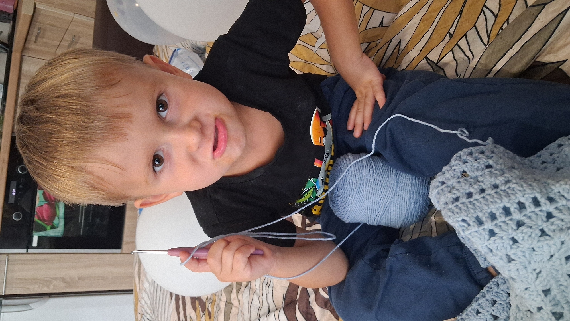 A young boy with blonde hair and brown eyes, wearing a black t-shirt with a colorful graphic and dark blue pants, sits on a bed with a patterned comforter. He holds a purple crochet hook with light blue yarn in his right hand. A large ball of light blue yarn is in front of him, and a partially crocheted blue item rests on his lap. The boy is looking directly at the camera with a slightly displeased expression, his mouth in a pout. In the background, kitchen cabinets and a stove are visible, suggesting the scene is likely within a residential setting in Koltsovo, Russia. Two white balloons are also visible in the background.