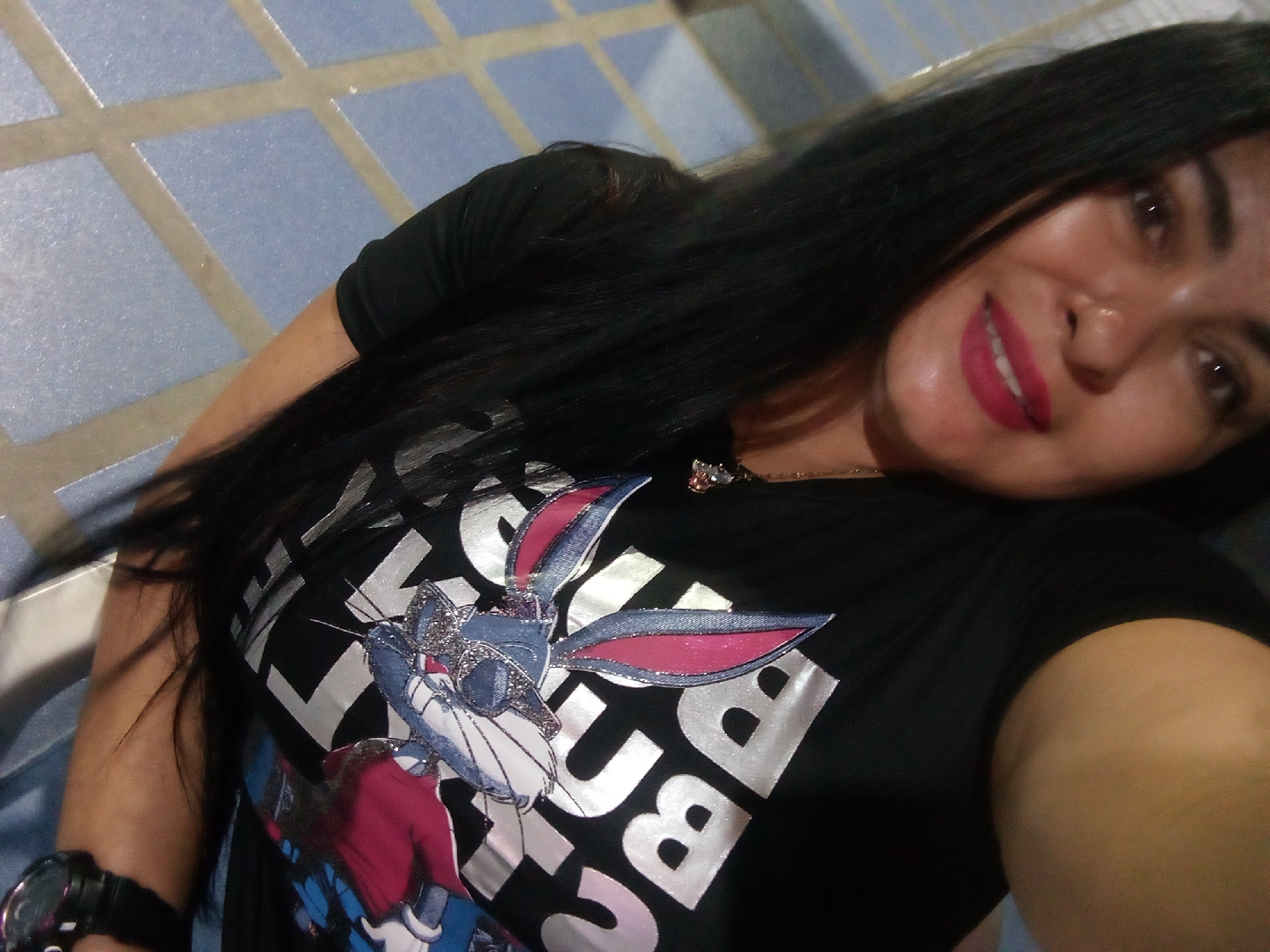 A medium shot captures a woman with long, dark hair, wearing a black t-shirt featuring a graphic of Bugs Bunny with silver lettering. She has fair skin and is wearing red lipstick. A watch is visible on her left wrist. The background appears to be a tiled surface, possibly a floor or wall, with a light blue and white pattern. The location is Unknown, Unknown.