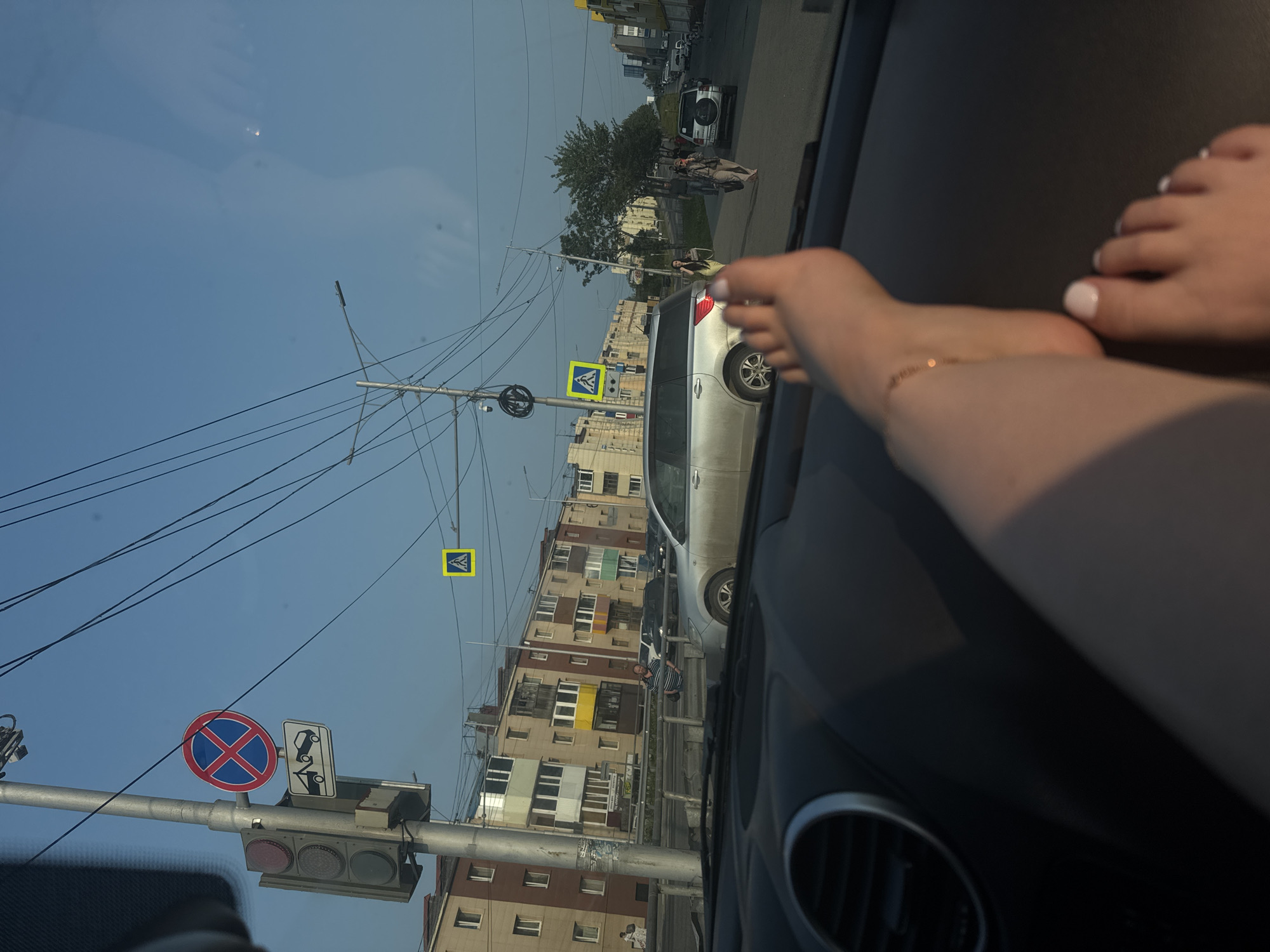 The image depicts a street scene in Petropavlovsk-Kamchatsky, Russia, viewed from the interior of a car. In the foreground, a person's bare feet rest on the dashboard, with one foot adorned with a thin gold anklet and the other showing white-painted toenails. The dashboard itself is dark and textured, with a visible air vent on the left.
Outside the car, the environment is a city street with buildings lining one side. These buildings are multi-story, with visible balconies and varying color schemes, including beige, brown, yellow, and green. A traffic light with red, yellow, and green signals is mounted on a pole, accompanied by a "No Parking" sign with a red circle and crossbar, and a sign depicting a tow truck. Further down the street, a pedestrian crossing is indicated by a yellow square sign with a white pedestrian silhouette.
Several vehicles are present, including a silver SUV, a black car, and a white SUV. Pedestrians are also visible on the sidewalk and crossing the street. The sky is clear and blue, suggesting daylight. The overall impression is a snapshot of everyday life on a street in a Russian city.