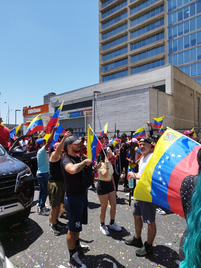 A crowd waves Venezuelan flags on a city street with confetti scattered about. People stand near a black SUV, a mix of men and women, some wearing hats or caps. A tall building looms in the background, adding to the urban setting. The atmosphere appears celebratory, suggesting a parade or rally. A sign for "Banco Consorcio" is visible in the background.