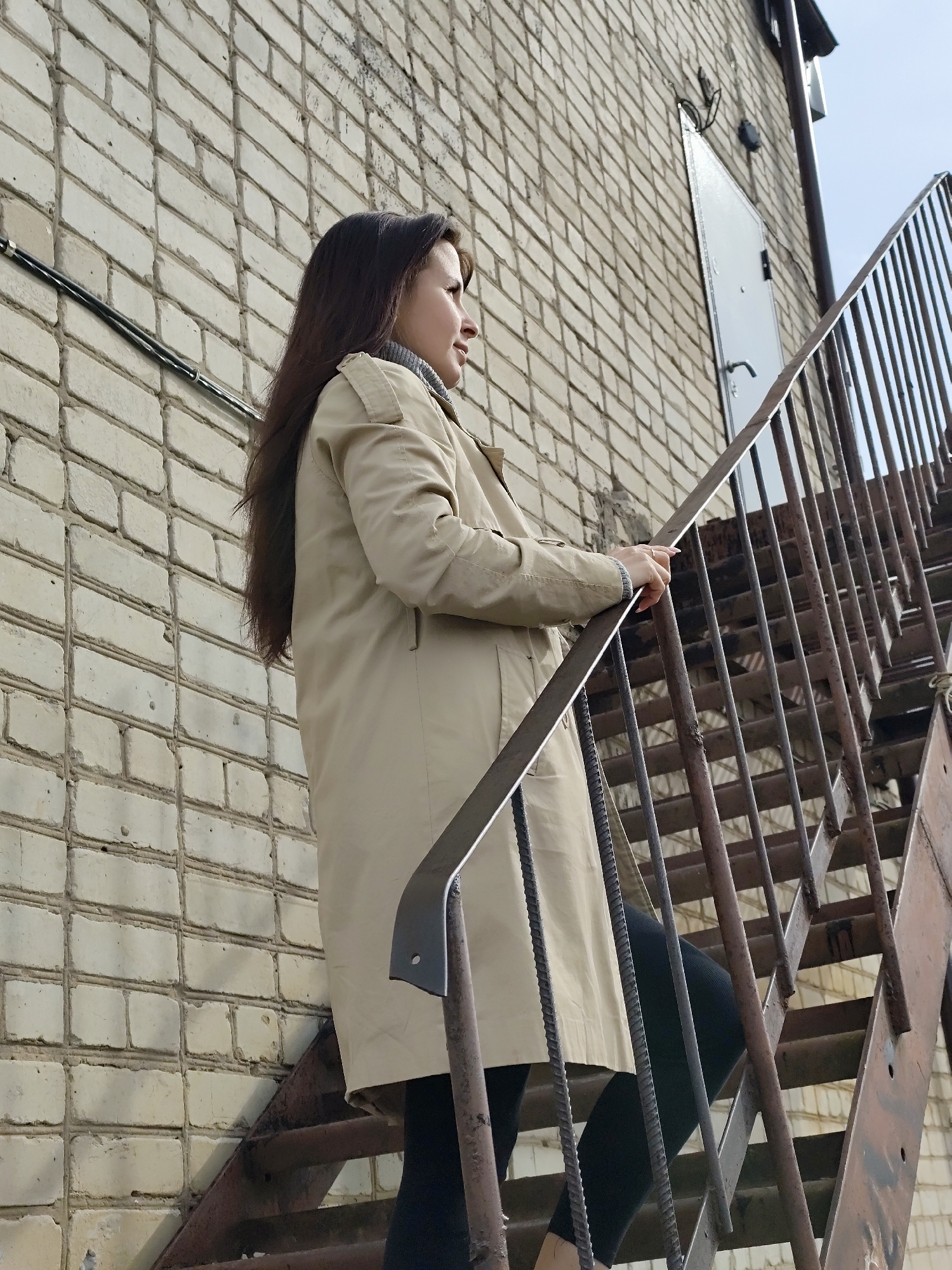 A low-angle, full-length shot captures a woman on an outdoor metal staircase, framed against a light-colored brick wall. The woman, with long dark hair, is seen in profile from the waist up, facing to the right and looking into the distance with a thoughtful or observant expression. She is wearing a beige trench coat over a grey turtleneck and dark leggings. Her right hand rests lightly on the railing of the rusty metal staircase as she appears to be ascending or pausing on the steps.
The background consists of a weathered, off-white or light-colored brick wall, showing distinct mortar lines. A black electrical conduit runs horizontally along the lower part of the wall behind her head. Higher up on the wall, to the right of the woman, a utilitarian white metal door with a handle is visible, set into the brickwork. Above the door, some wires and fixtures are attached to the wall. The metal staircase itself has an industrial look, appearing worn and possibly rusty, with simple vertical bar railings and open risers. A patch of clear sky is visible in the upper right corner. The lighting suggests it is daytime, providing ambient illumination without harsh shadows. The overall setting implies an urban or industrial area in Фоки, Russia. There is no visible text in the image.
