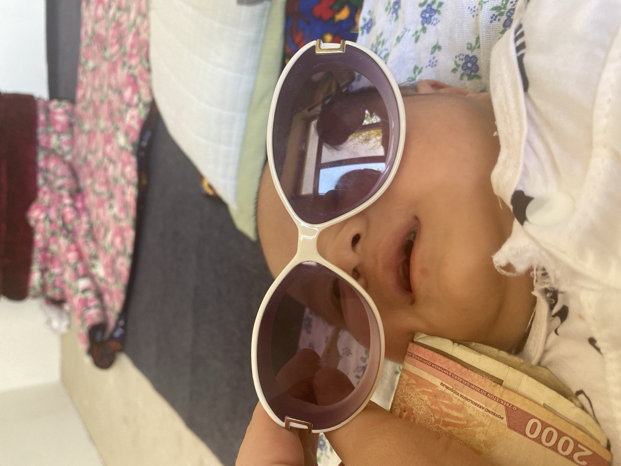 In Arbagish, Uzbekistan, a close-up shot shows a baby's face partially obscured by large, white-framed sunglasses with purple lenses. A hand holds the sunglasses, and a stack of Uzbekistani 2000 Som banknotes is placed near the baby's cheek. The background includes blurred floral-patterned fabric and bedding.