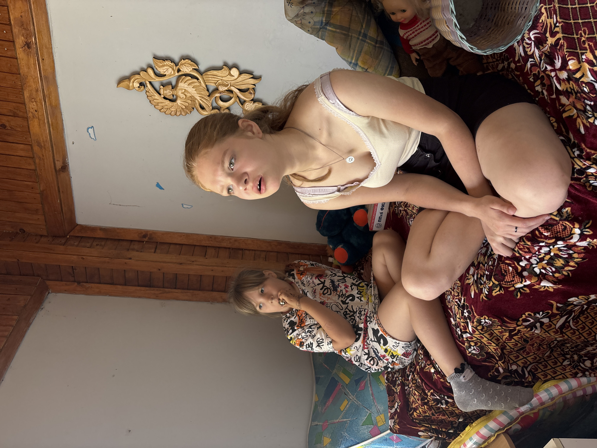 The image depicts an indoor scene with two young girls seated on a patterned blanket spread across a low surface, possibly a bed or sofa. The girl on the left, with blonde hair and bangs, wears a white dress adorned with colorful graffiti-style text and illustrations. She is holding something to her mouth and appears to be eating or putting something in her mouth. Her left leg is crossed over her right, and she wears a grey sock with a black design on her left foot. The girl on the right, also with blonde hair tied back in a ponytail, is wearing a cream-colored tank top with a lace trim and black shorts. She is looking upwards and to the left with an open mouth, as if speaking or reacting to something. Her legs are crossed, and she is wearing a necklace with a circular pendant.
The background features a wall painted a light gray, with wooden paneling and beams in the upper portion of the image. A carved wooden ornament, possibly depicting a bird-like creature with a human face, is mounted on the wall above and to the right of the girl on the right. To the right of this ornament, a pillow with a plaid pattern is visible, and next to it, a doll and a woven basket. To the left of the girls, a bolster or pillow with a geometric pattern is partially visible. The overall environment suggests a rustic or simple living space. The presence of wooden elements and the style of the furniture and wall decorations hint at a location in a more rural or traditional setting. The question asks for the location in the city of Аряж, Russia, but there is no visual information in the image that can confirm or deny this specific location.