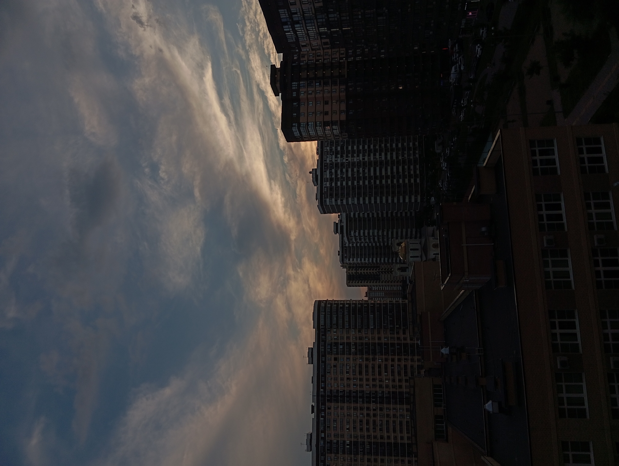 The image depicts a twilight view of a residential area in Krasnodar, Russia. The sky is filled with dramatic clouds, painted in hues of grey, blue, and a soft orange glow from the setting sun. Dominating the scene are several multi-story apartment buildings, characteristic of modern urban architecture. The buildings vary in design and color, with some featuring lighter facades and balconies, while others are darker and more monolithic. In the distance, a golden dome of a church or religious building is visible peeking above the rooftops. Below the apartment buildings, a street with parked cars and some greenery can be observed, suggesting a typical urban neighborhood. The overall atmosphere is tranquil and serene, capturing the transition from day to night in a Russian city.