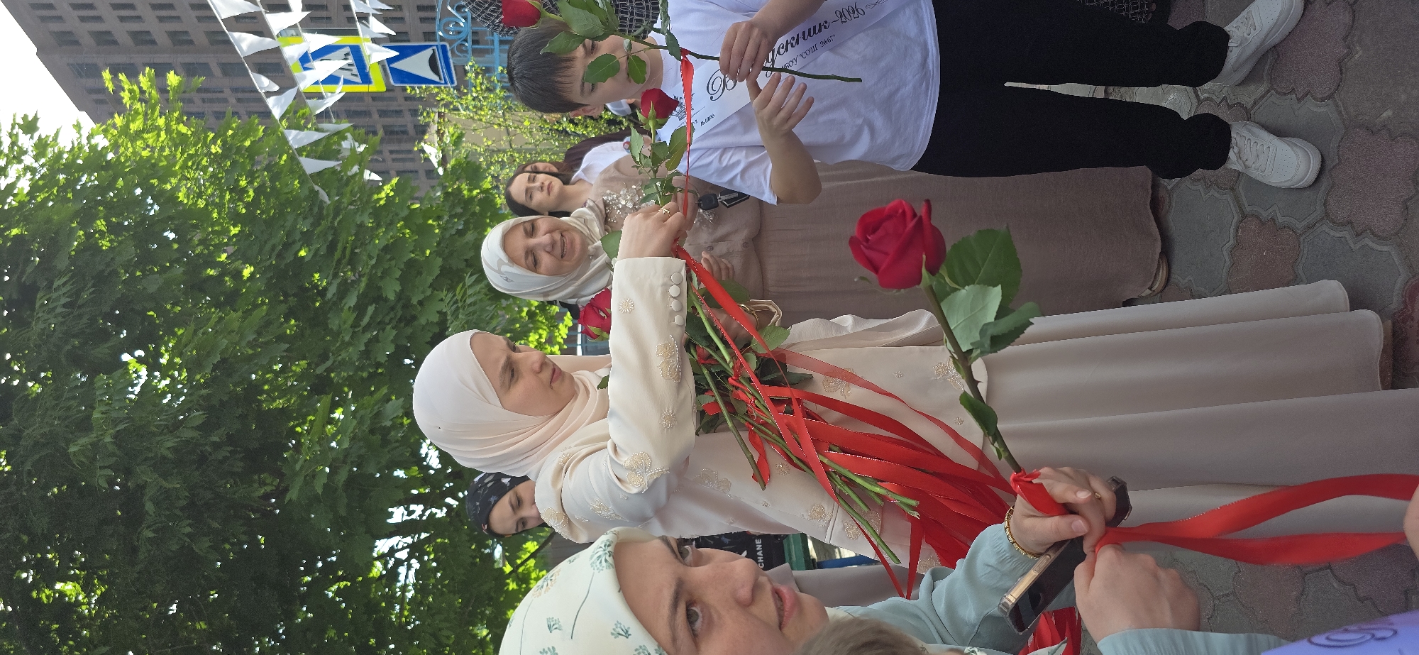 A group of people stand in an urban environment, holding red roses. Most of the individuals are women wearing headscarves, while at least one young male is present. A woman in a light-colored outfit stands at the forefront, clutching several roses tied with red ribbons. Other members of the group, also in traditional clothing, look on, some holding roses as well. A young man in a white t-shirt is also seen holding a rose.
The background includes a building with multiple windows and foliage, suggesting a park or tree-lined street. Above, there is a series of white triangular flags stretched overhead, indicating an event or celebration. The ground appears to be paved, with patterns visible on the surface.
The location is described only as Unknown, Unknown.