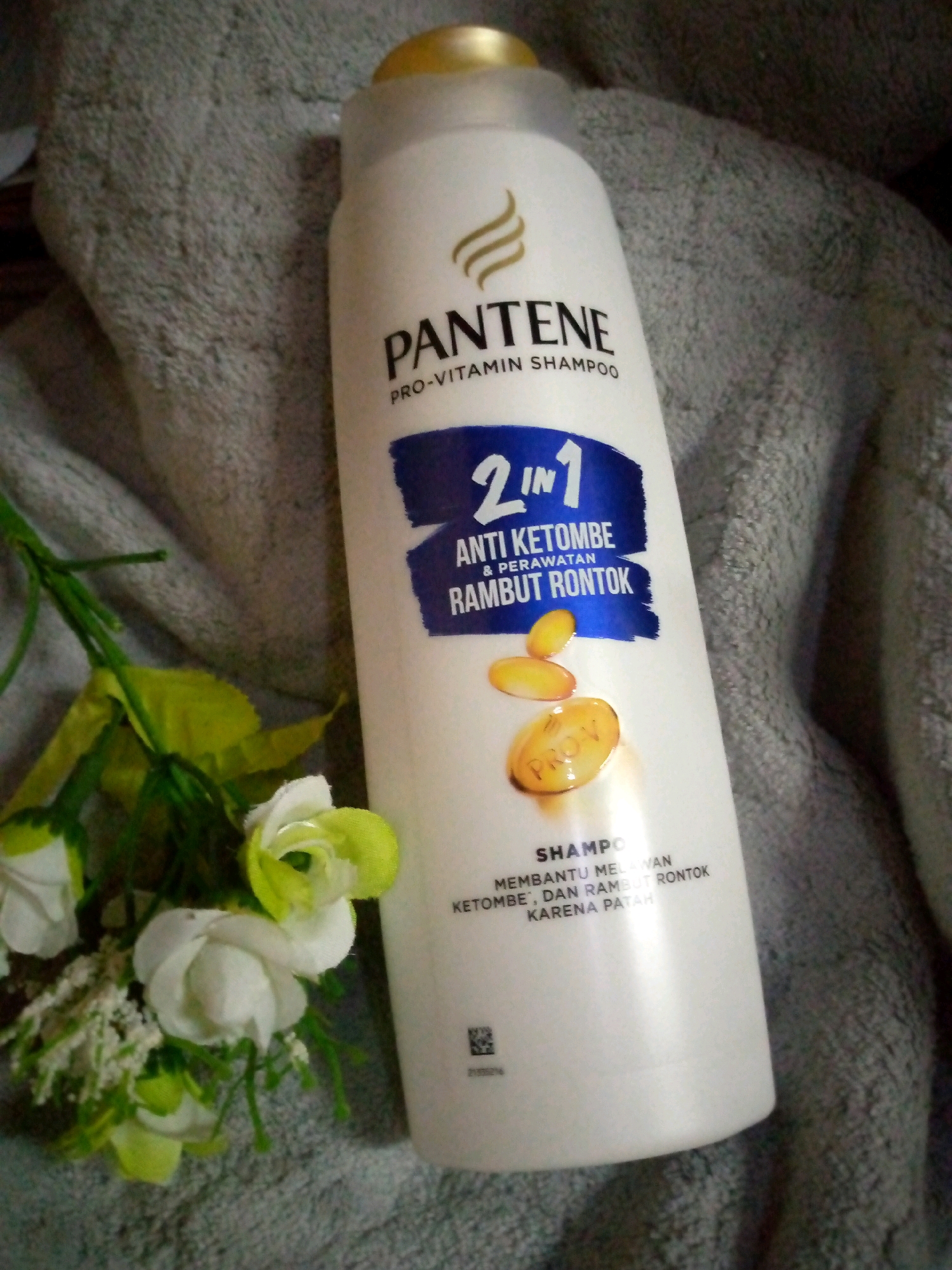 A white Pantene Pro-Vitamin Shampoo bottle, featuring a gold cap, is positioned centrally on a gray textured surface, likely a towel. The bottle displays a blue label with the text "2 IN 1 ANTI KETOMBE & PERAWATAN RAMBUT RONTOK," and depicts three yellow oval capsules. To the left of the bottle, a small arrangement of artificial flowers, composed of white and light green blossoms and green leaves, is visible. The scene is well-lit, highlighting the product and its surroundings. The image is a product shot, with no indication of any specific location in Sumberwono, Indonesia.