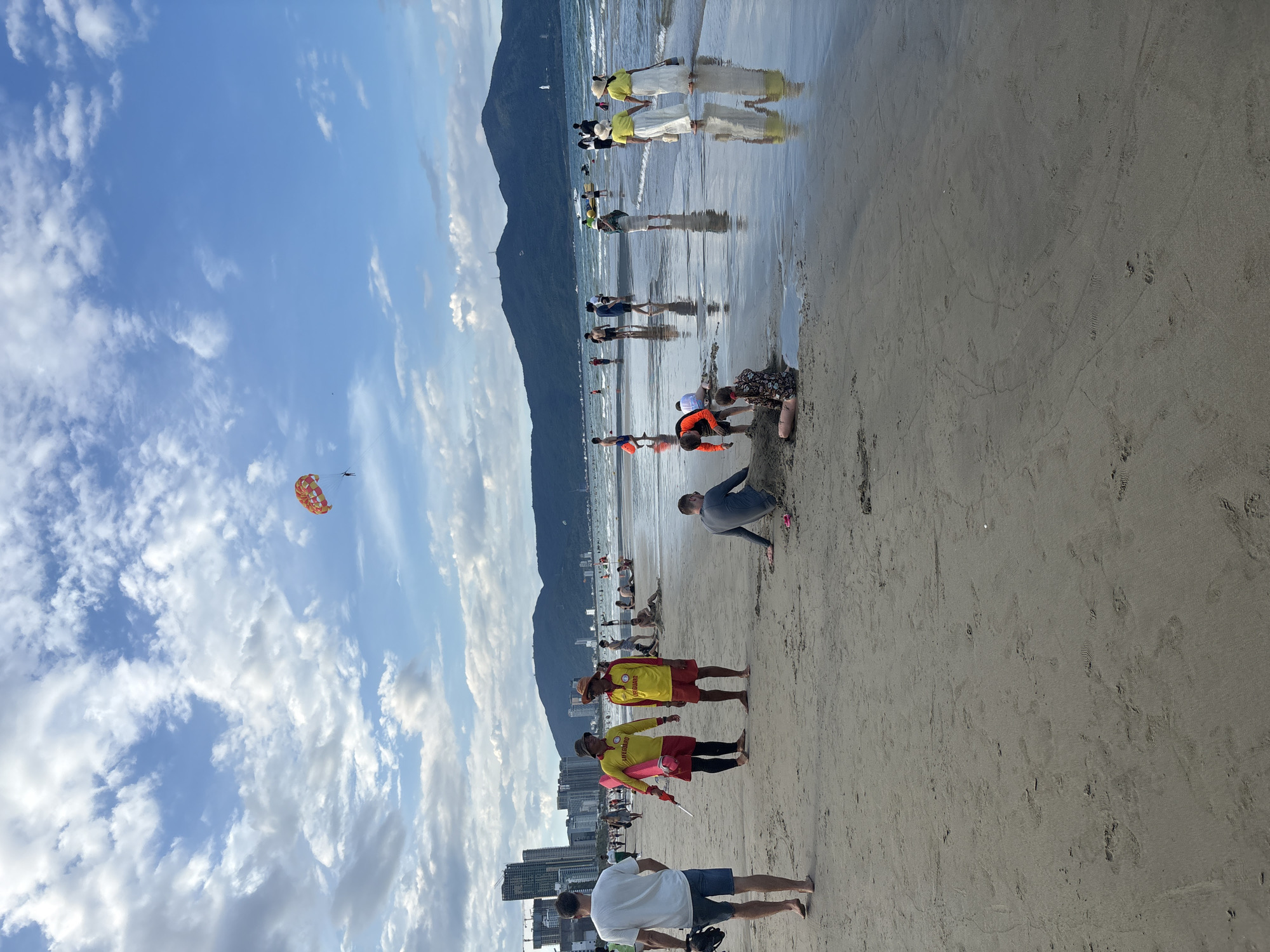 The image depicts a lively beach scene on a sunny day in Da Nang, Vietnam. The beach is wide and sandy, with the gentle waves of the sea lapping at the shore. In the foreground, several people are enjoying various beach activities. Two men in red shorts and yellow "LIFEGUARD" shirts are standing and conversing, one holding a rescue buoy. Nearby, a man in a grey shirt is seated on the sand, seemingly engaged in building a sandcastle with another person. Further down the beach, a group of people are walking near the water's edge, some holding hands and others carrying bags. Children are playing in the shallow water and sand.
In the mid-ground, the ocean is filled with activity. Several boats and jet skis are visible, and a person is parasailing in the sky, pulled by a boat. The coastline is lined with modern high-rise buildings, indicating the urban development of Da Nang. In the background, lush green mountains form a picturesque backdrop to the scene. The sky is a vibrant blue, dotted with fluffy white clouds, adding to the overall pleasant atmosphere. The reflections of the people and the sky on the wet sand create a beautiful, shimmering effect. The image captures a typical day at a popular tourist destination, with a blend of relaxation, recreation, and natural beauty.