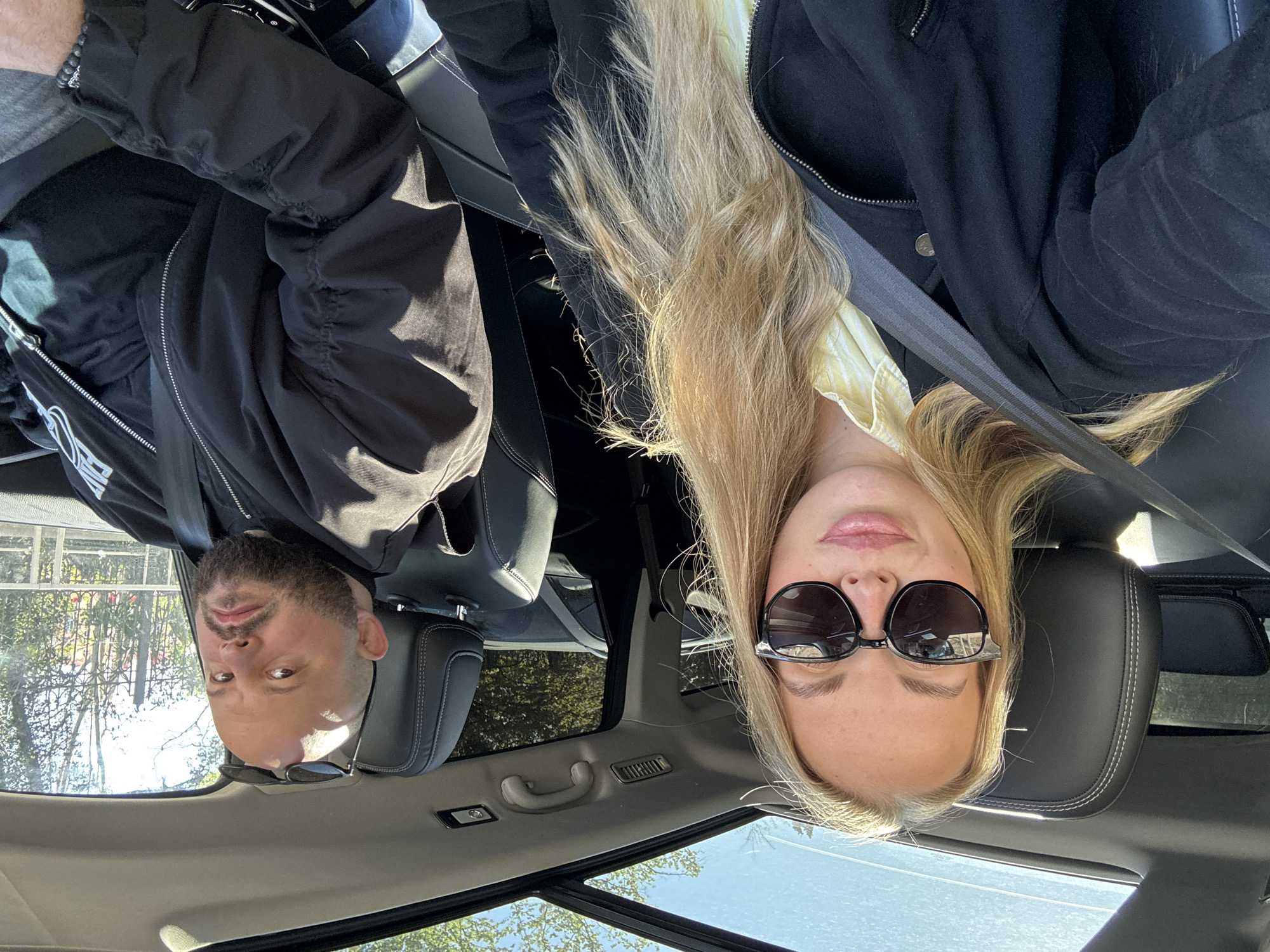 The image, intentionally rotated 180 degrees, captures two individuals inside a car in Sochi, Russia, on a bright, sunny day.
After mentally rotating the image, a male subject is visible on the left side, characterized by short dark hair and a beard. He wears a black jacket over a grey t-shirt, with black sunglasses resting on his nose. A white, stylized logo, possibly reading "PIN", is partially visible on his jacket. His expression is neutral.
On the right side, a female subject with long, straight blonde hair is present. She wears black sunglasses and a black jacket over a light yellow or cream-colored top. Her expression is also neutral, showing a slight hint of a smile.
Both individuals are seated in black leather car seats and are properly secured with seatbelts, suggesting they are either driving or passengers. The car interior features black upholstery and a ceiling handle. Above them, a panoramic roof or sunroof reveals a clear blue sky and the tops of green trees, indicating an outdoor setting. The bright light and visible shadows confirm it is daytime with sunny weather. The image appears to be a selfie taken from a low angle, capturing both subjects looking towards the camera in a relaxed manner.