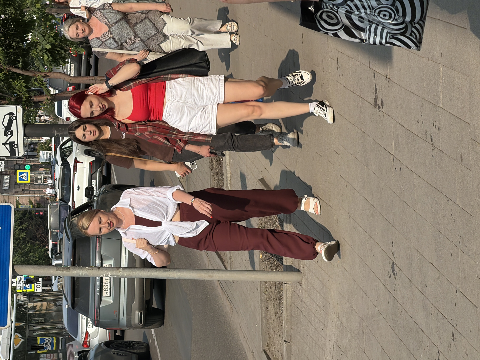 The image captures a street scene in Krasnoyarsk, Russia, likely on a sunny day, as indicated by the strong shadows. Four women are the central subjects, walking along a paved sidewalk. The woman in the foreground on the left is wearing a white open-collared shirt tied at the waist and maroon wide-leg trousers, paired with white sandals. She is holding a light-colored purse and has her right hand raised. To her right, a woman with red hair is dressed in a red tank top, white shorts, and a plaid shirt, wearing black and white sneakers. She is adjusting an earbud. Behind her and slightly to the left is a woman with dark hair in a brown t-shirt and dark pants, also wearing sneakers. On the far right, an older woman in a patterned blouse and light-colored trousers walks with a tote bag.
The environment includes a street with several cars, including a gray SUV with the license plate "H836T" visible. There are traffic signs and streetlights, and a metal pole stands prominently in the foreground, partially obscuring the view of the woman in maroon. Trees and urban buildings form the backdrop. The perspective is from a slightly elevated viewpoint, looking down at the sidewalk and street.