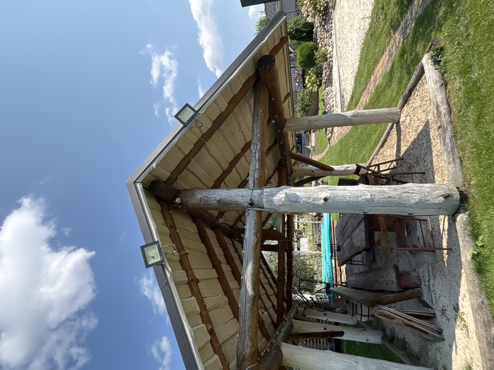 This image depicts an outdoor wooden gazebo with a rustic design, likely located in Zvenigorod, Russia. The gazebo features a sloped roof supported by thick, natural log posts. The roof is covered with light-colored tiles and has two modern floodlights mounted on its exterior. Inside, a large, rough-hewn wooden table is set with wooden benches and chairs. To the left, a wooden barrel and folded wooden items are visible. In the background, a bright blue swimming pool is partially visible, surrounded by a sandy area and lush green grass. The environment suggests a recreational or leisure space, possibly a backyard or a resort area, with a clear blue sky dotted with white clouds overhead.