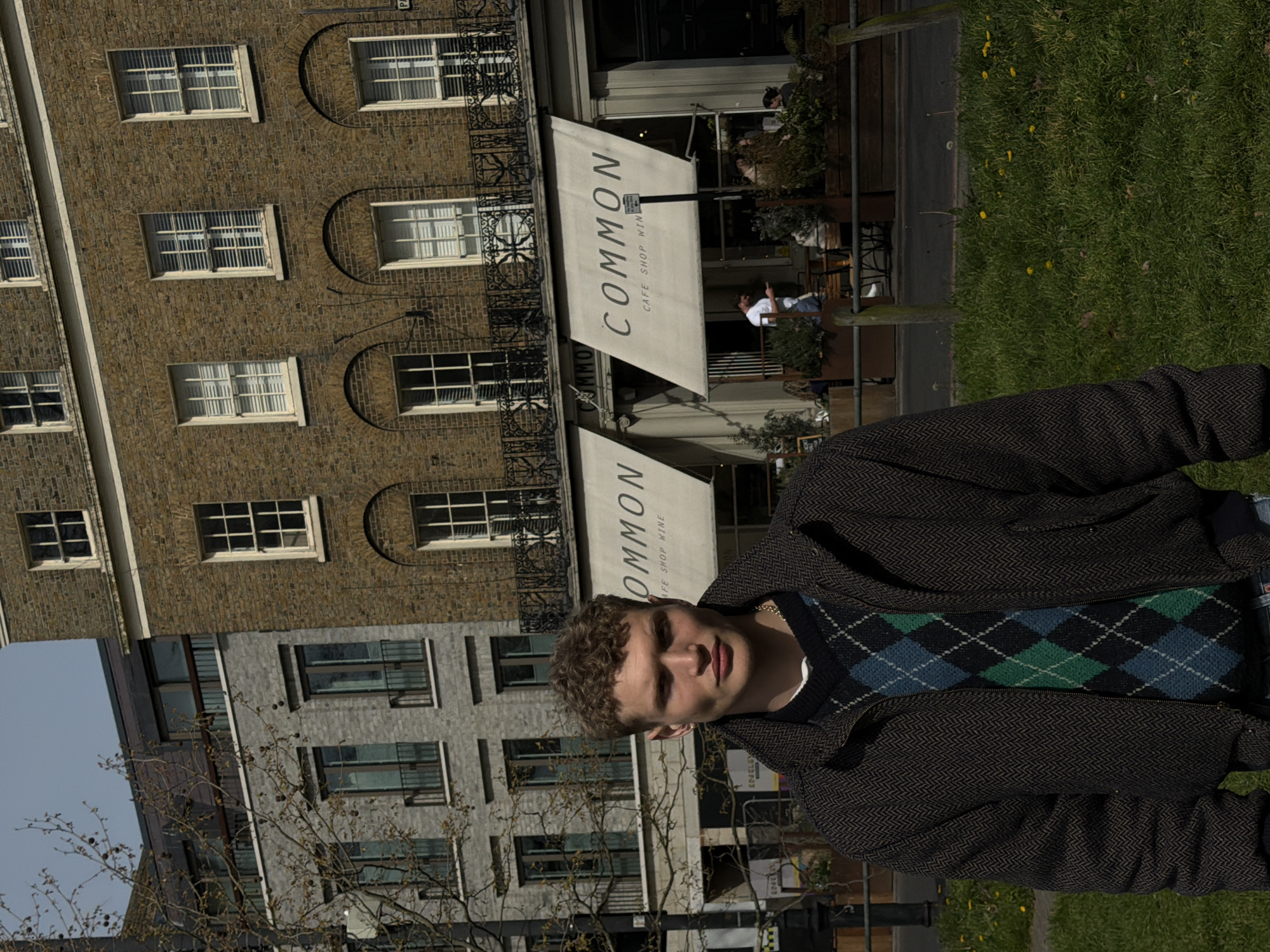 A young man with curly blonde hair stands outside a building labeled "COMMON CAFE SHOP WINE" in London, United Kingdom. He wears a patterned sweater and dark-colored jacket. The building behind him has rows of windows and is located near a grassy area. A second building of light gray/silver color is visible in the background.