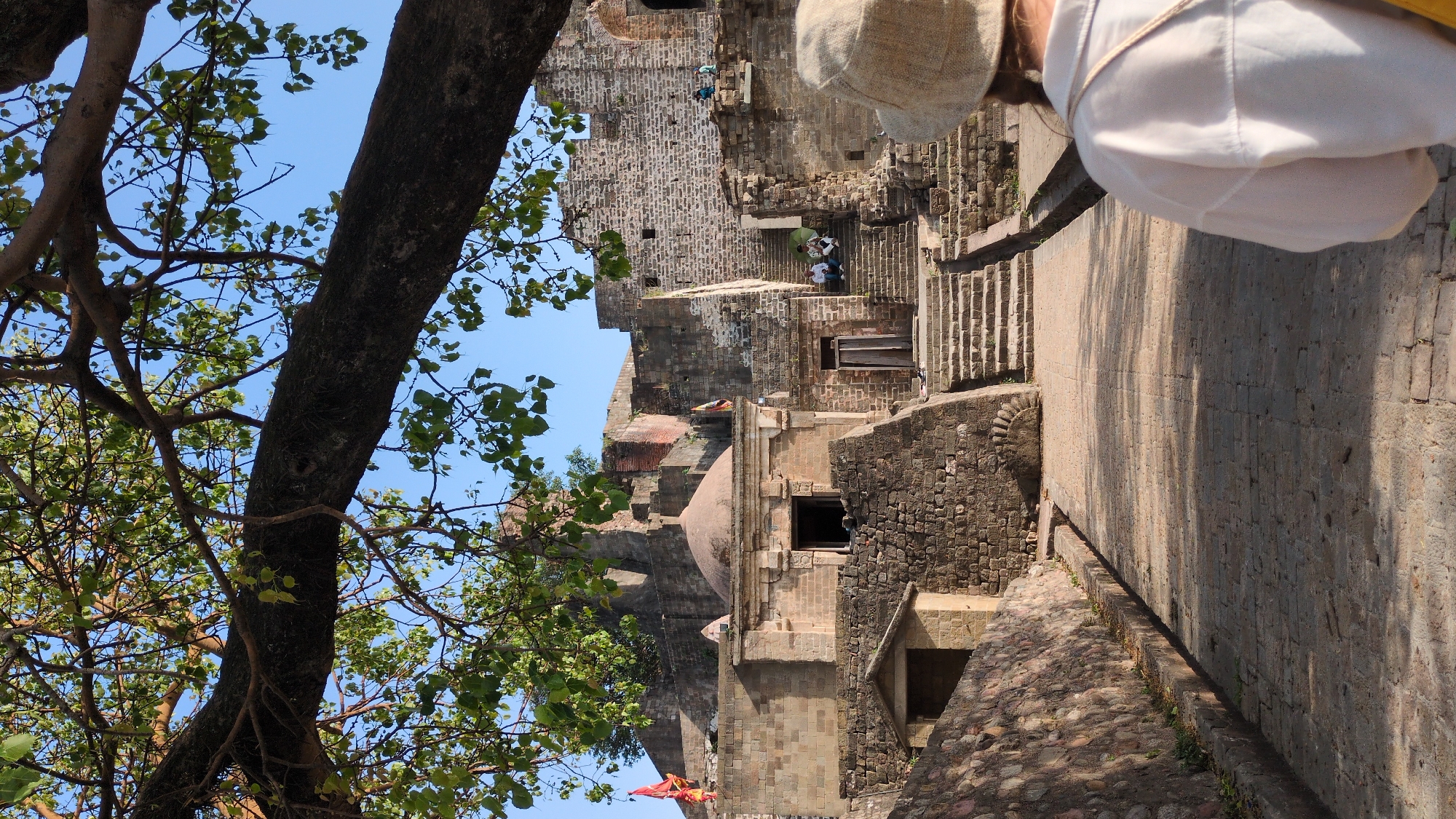 The image shows an outdoor scene in Kaswara, India, likely a historical or tourist site. The scene includes stone structures, potentially part of a fort or ancient complex. In the foreground, a large tree with a thick trunk and leaves occupies the left side of the frame, partially obscuring the structures behind it. The stone buildings exhibit different architectural styles, including a dome and arched openings. In the middle ground, a person with a hat and white clothing is positioned on the right side, with stone walls and steps visible. The environment suggests a dry, sunny day. People are seen in the background walking on the stone steps.