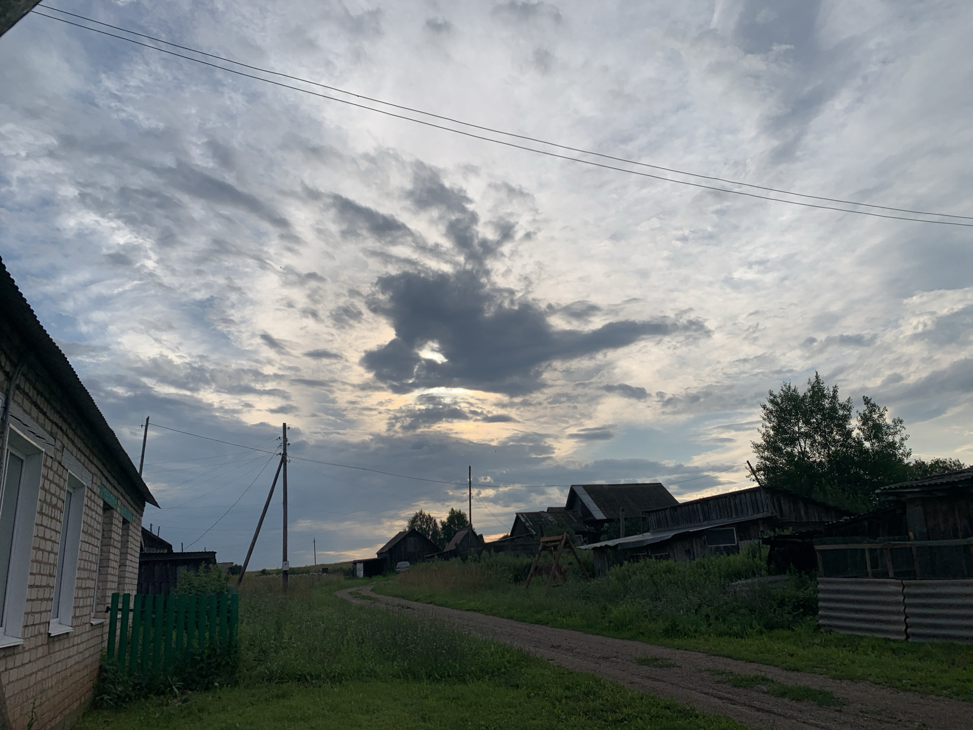 This is a medium-angle, eye-level, outdoor, horizontal, daytime shot of a rural scene in Panteleevka, Russia, under a cloudy sky. In the foreground to the left is the side of a light-brown brick house with white-trimmed windows. A short, green, wooden fence is in front of the house. A dirt path curves from the foreground towards the background and disappears between buildings. The background features a collection of dark, weathered wooden structures, some with corrugated metal roofs. Tall grass and weeds grow throughout the scene. Power lines and utility poles are visible in the midground and background. The sky is filled with a variety of grey and white clouds, with a patch of brighter light peeking through in the center, suggesting either sunrise or sunset. No people are visible in the image.