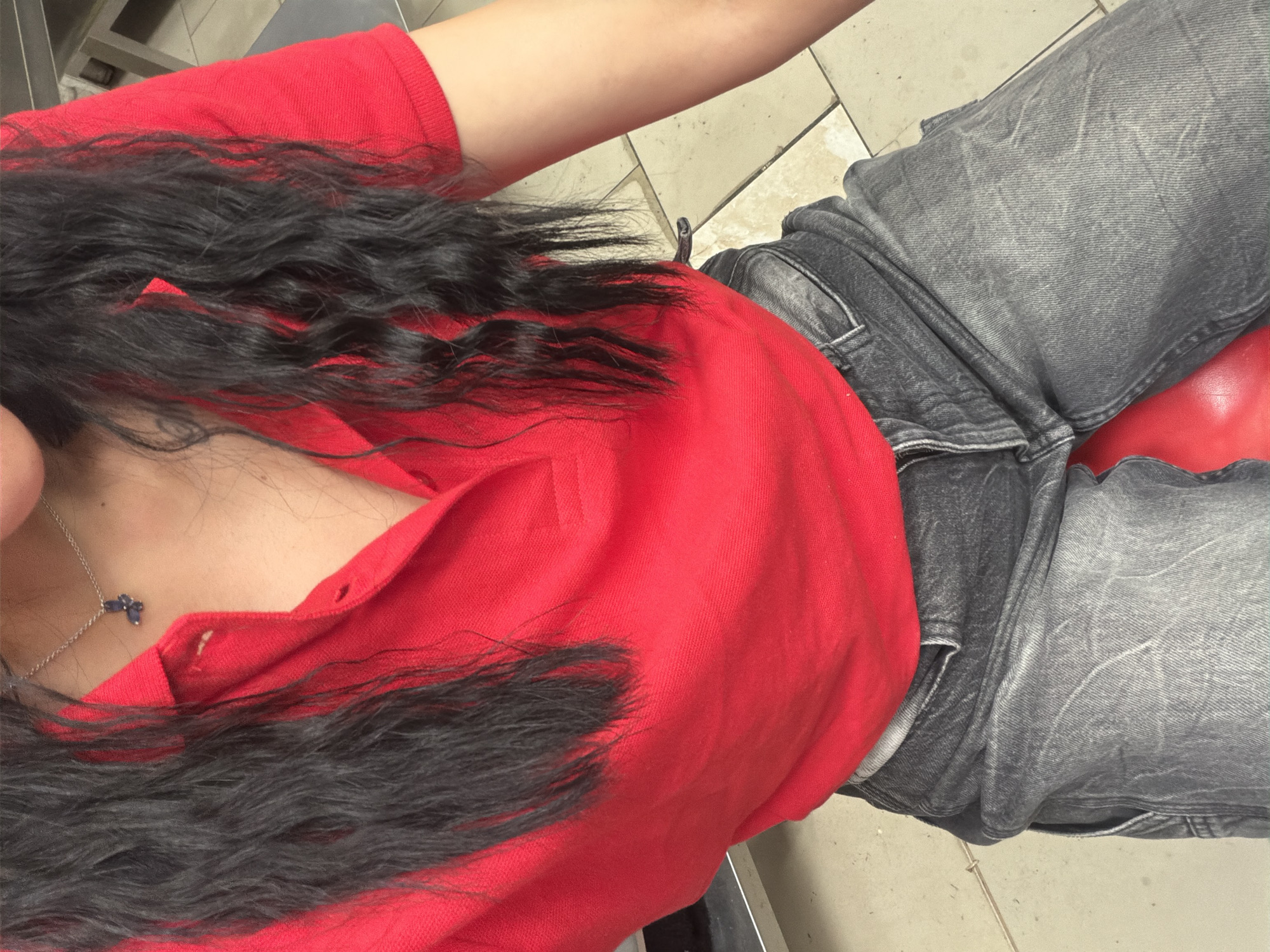 A close-up shot shows a person from the chest down, sitting. The person is wearing a bright red collared shirt and dark gray distressed jeans. A silver necklace with a small, dark blue pendant is visible on the person's neck. Long, dark, wavy hair cascades over the red shirt and onto the jeans. The background consists of light-colored tiled flooring and a hint of a gray surface, possibly a chair or furniture. The lighting suggests an indoor environment, with no obvious cues for time of day or weather. The scene is static, implying the person is simply sitting. There is no visible text in the image. The location context is Plovdiv, Bulgaria.