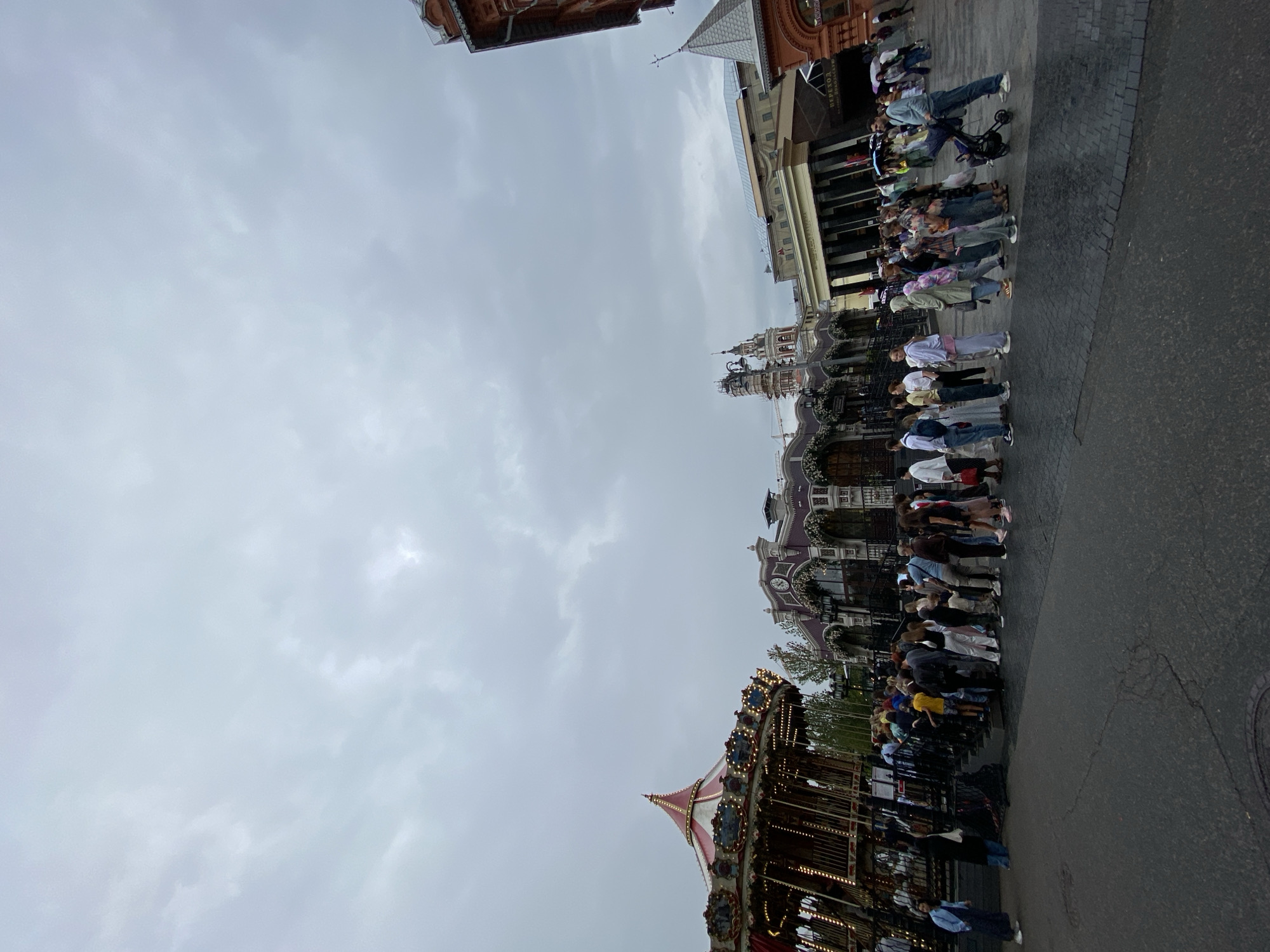 The image depicts a scene in Moscow, Russia, specifically Red Square, on an overcast day. A large crowd of people is gathered, forming queues near a carousel and several ornate buildings. The carousel, adorned with lights and red and white striped canopy, is positioned on the left side of the frame. To the right of the carousel, a detailed building with arched windows and floral decorations is visible. Further in the background, distinct architecture with towers and columns characteristic of Moscow's historical buildings can be seen. People of various ages are dressed in casual attire, with some holding umbrellas, suggesting potential rain. The ground is paved with cobblestones. The overall atmosphere suggests a public gathering or event taking place in a tourist area.