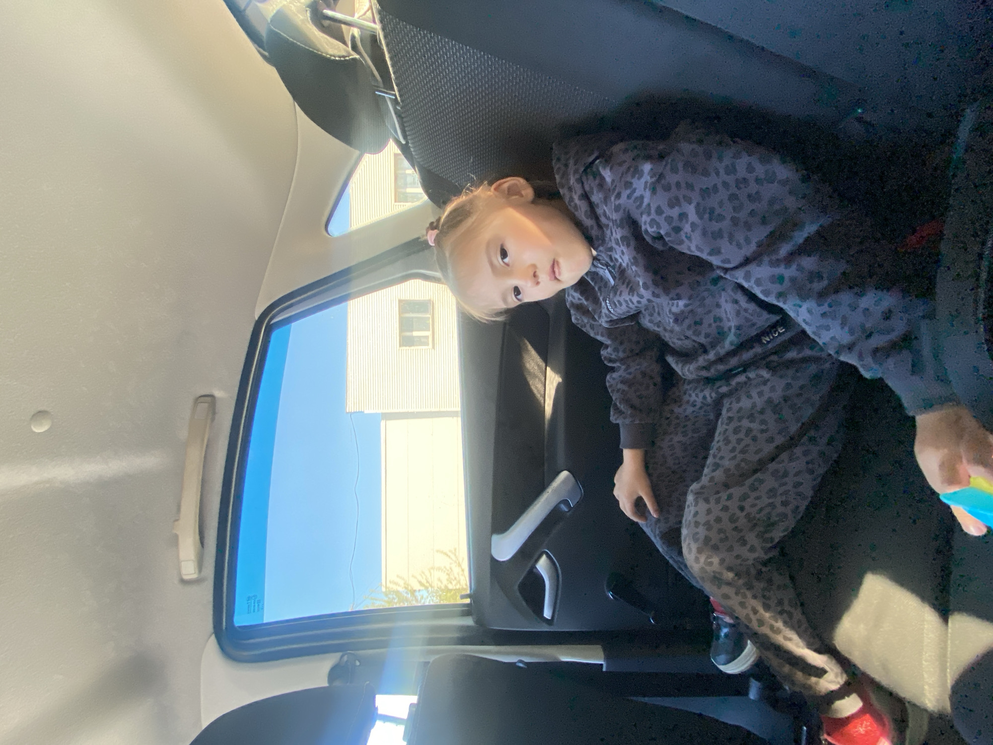 A young girl is sitting in the back seat of a car. She is wearing a grey leopard-print tracksuit and has pigtails. She is looking towards the camera. The car window shows a building with multiple windows, located in Chaikovsky, Russia. The sky is blue.